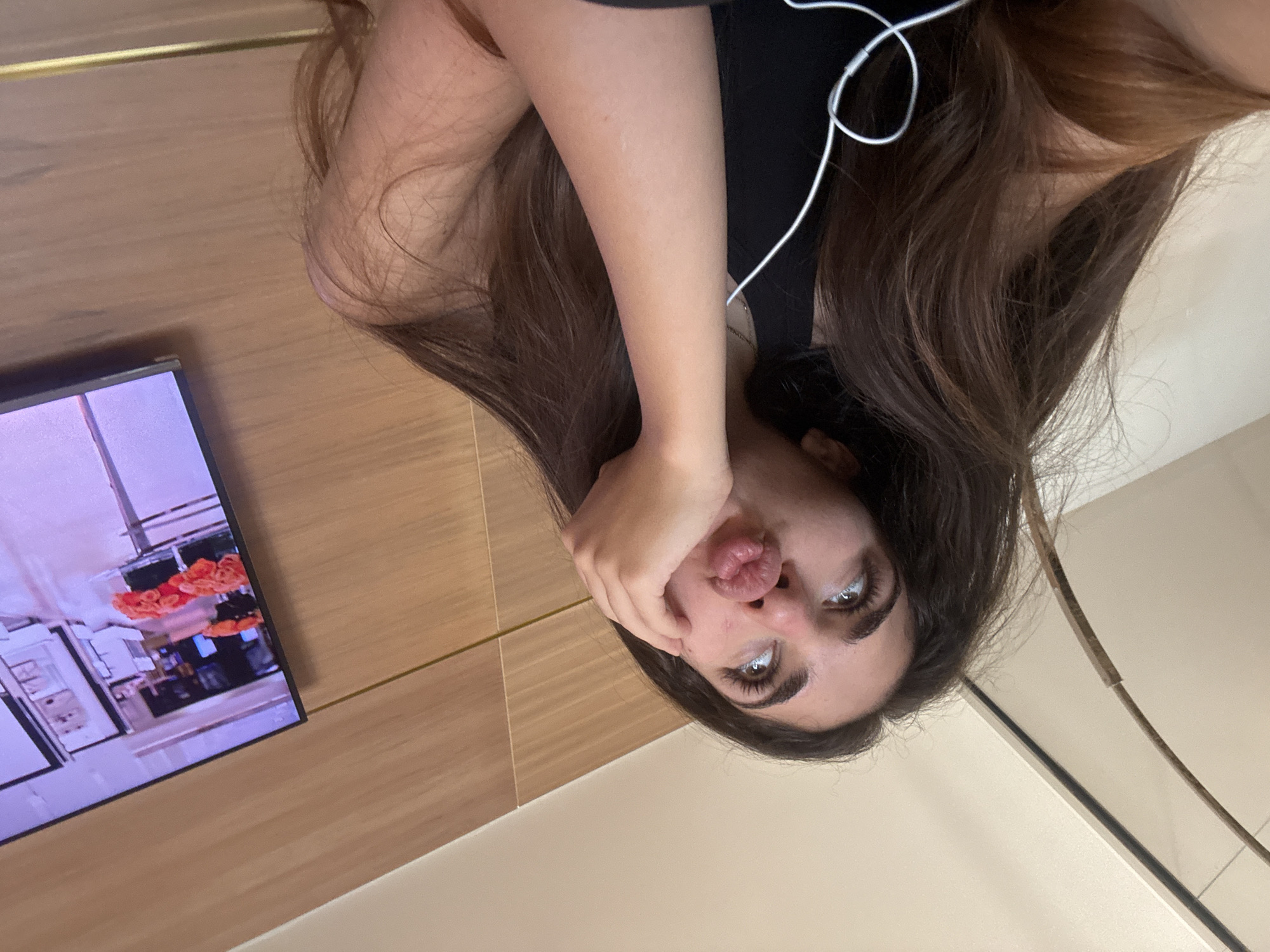 The image captures a close-up, low-angle shot of a young woman with long, dark brown hair, who appears to be in a relaxed or bored posture. Her head is tilted upwards, and her eyes are looking towards the ceiling. Her left hand is resting on her cheek, with her fingers extended towards her temple, and her lips are puckered in a pout. She is wearing a black tank top with white earbuds visible around her neck. In the background, to the right, a television screen displays an image of several bouquets of orange roses, suggesting an indoor setting. The wall behind the woman and the television is a wood-paneled surface with horizontal lines. The ceiling is a plain, light-colored surface with a curved element visible on the left side. The overall impression is a candid moment, possibly taken in a home or hotel room. The specific location in Boca Raton, United States of America, cannot be determined from the visual information provided in the image.