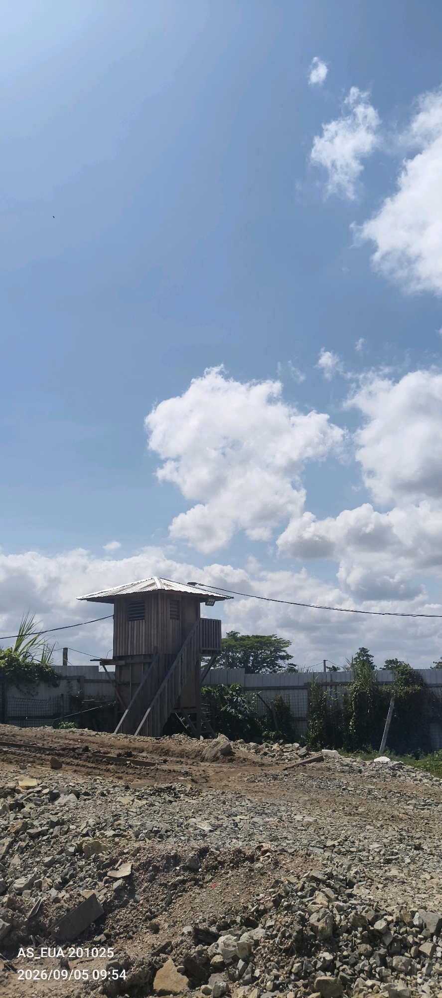 A vertical shot captures a small, wooden guard tower with a corrugated metal roof situated in a construction area. The tower has a platform and stairs leading up to it, with louvre windows on its sides. It is surrounded by a fence, with lush green vegetation and trees visible in the background under a bright blue sky dotted with fluffy white clouds. The foreground is filled with rough terrain, characterized by piles of rocks and dirt, suggesting ongoing construction or land clearing. A timestamp overlay at the bottom left of the image reads "AS_EUA 201025 2026/09/05 09:54". The location is identified as Kanduanum 2, Papua New Guinea. No people are visible in the image.