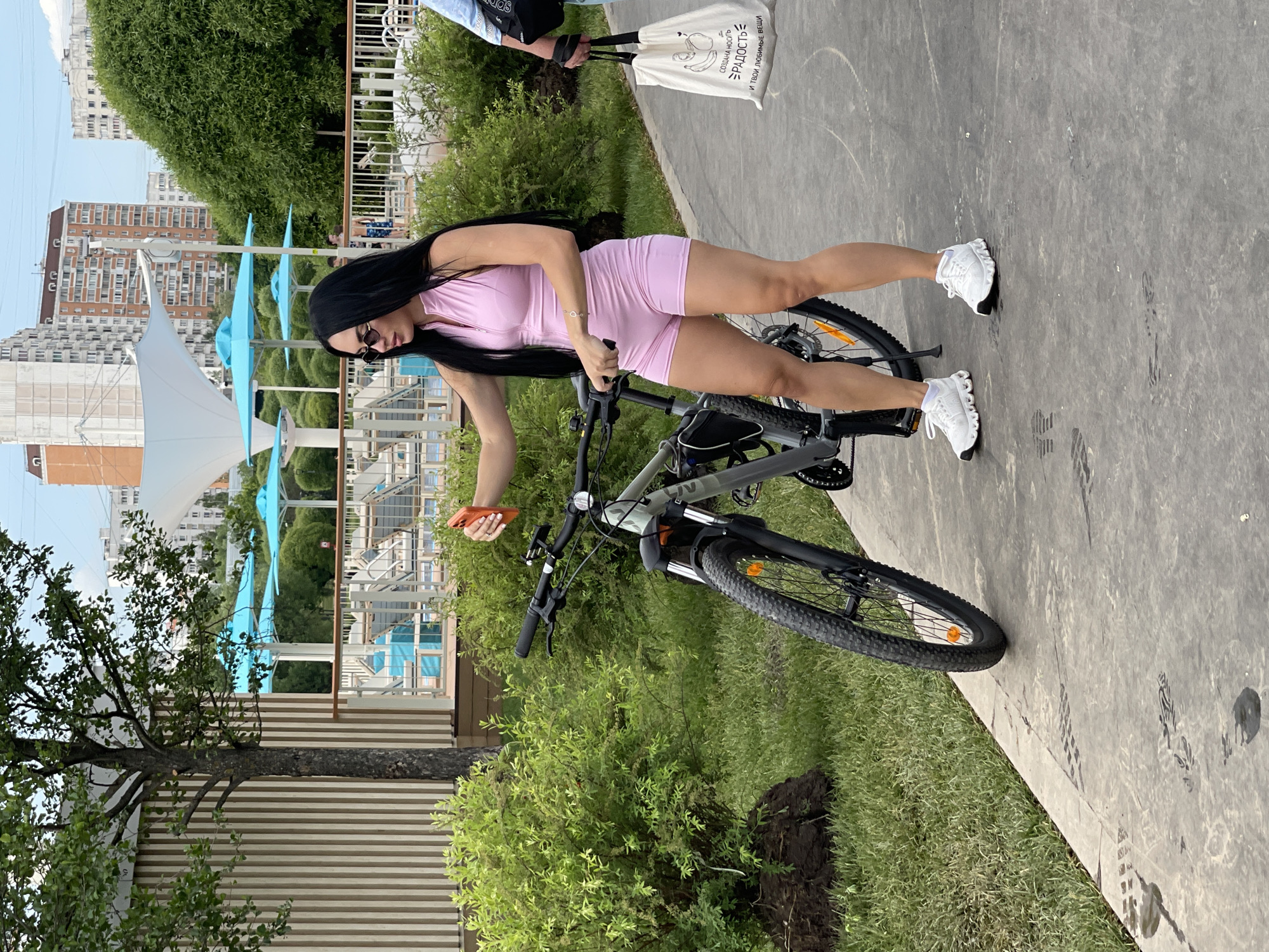 The image, taken in what appears to be Moscow, Russia, showcases a young woman standing on a paved path beside a bicycle. She has long, dark hair and is dressed in a light pink, form-fitting sleeveless romper with a front zipper, accessorized with sunglasses and a silver bracelet on her left wrist. She wears white athletic shoes and is holding an orange smartphone in her right hand, while her left hand rests on the bicycle's handlebars.
The bicycle is a dark-colored mountain bike with front suspension and an orange reflector on the front wheel. Surrounding the path are green bushes and trees. In the background, under a clear sky, are multi-story residential buildings, typical of a city environment. Closer in the middle ground, light blue sun umbrellas and white fabric structures suggest a recreational area or poolside setting.
To the right of the woman, another person is partially visible, holding a white canvas tote bag. The bag features a graphic of bananas and Russian text, which translates to: "CREATING NEW JOY AND YOUR FAVORITE THINGS." The scene depicts an outdoor setting on a bright, sunny day, with the woman seemingly pausing during an activity involving her bicycle.