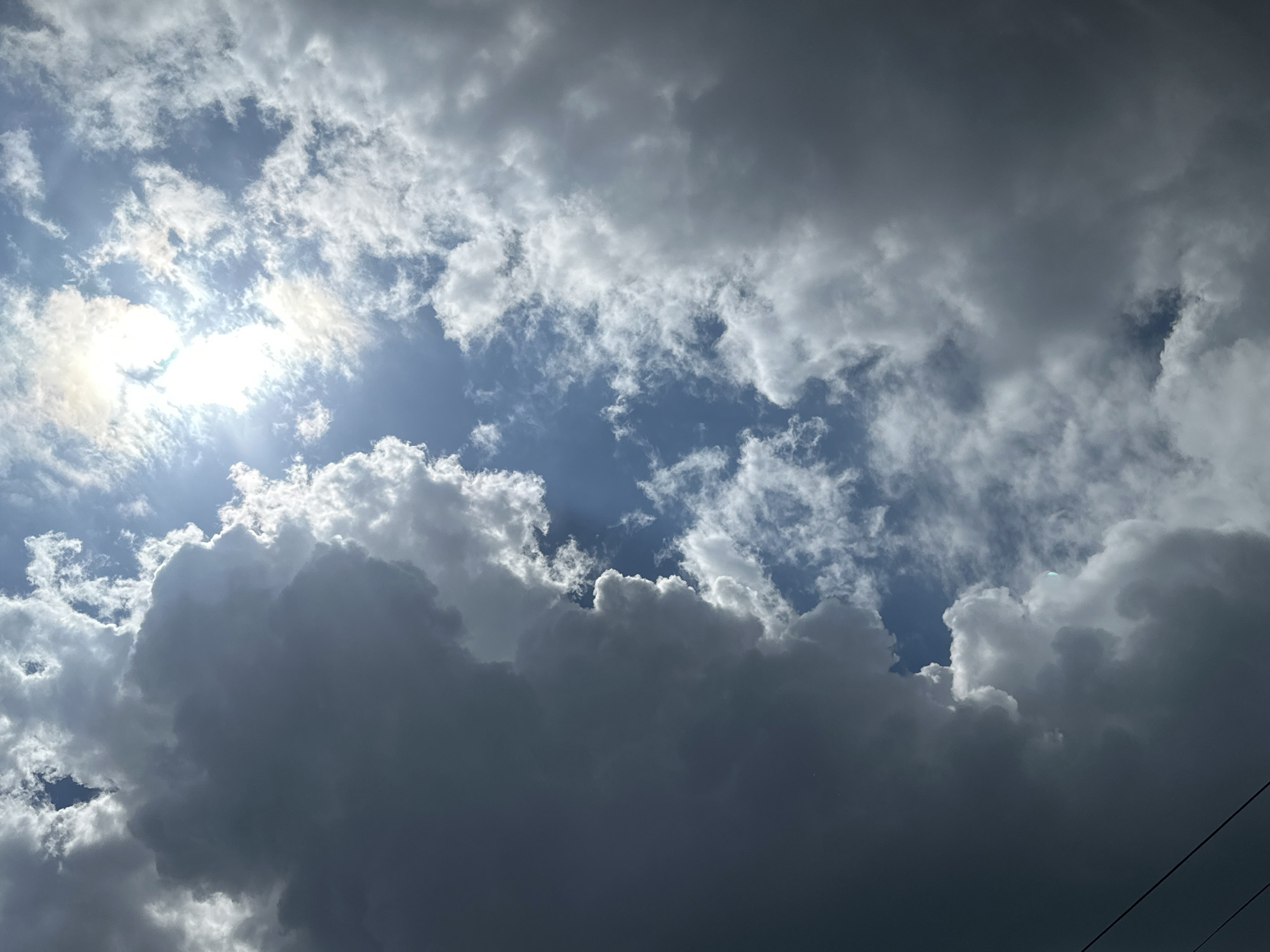 A low-angle shot captures a dynamic sky filled with a mix of dark, looming storm clouds and bright, sunlit patches. The sun is visible in the upper left quadrant, shining through thinner clouds with a brilliant white glow, casting rays that illuminate the edges of some clouds. The clouds themselves vary in density and color, ranging from deep grays and blues in the stormier areas to lighter whites where the sun's rays penetrate. The overall impression is one of impending weather change, with both darkness and light coexisting. The bottom right of the image shows two thin, dark power lines crossing diagonally. The scene is devoid of people or any man-made structures, suggesting an open sky environment.