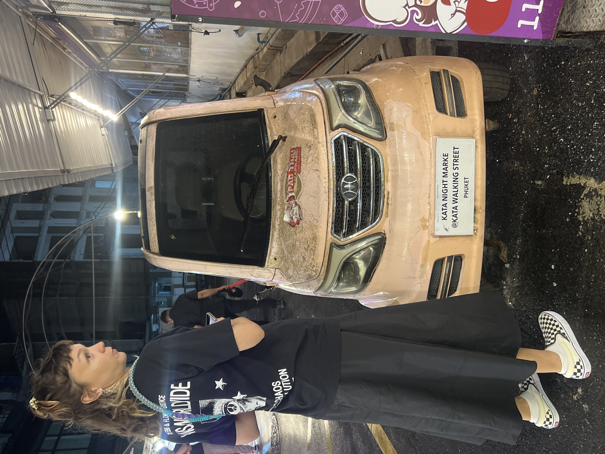 In a nighttime street scene in Karon, Thailand, a woman with wavy brown hair and bangs stands facing left, looking upwards. She is wearing a black t-shirt with white text and graphics, a long black skirt, and checkered Vans slip-on shoes. A beaded necklace with blue and purple accents is around her neck.
Beside her, a light pink vehicle, possibly a small car or a tuk-tuk, is parked on a dark, wet asphalt surface. The vehicle has a slightly worn appearance, with some peeling paint. A sticker on the front reads "PAD THAI MAPRAN" and depicts a cartoon character. Another sign on the vehicle's bumper says "KATA NIGHT MARKE @KATA WALKING STREET PHUKET".
In the background, there are market stalls with metal roofing and lights, suggesting a bustling night market atmosphere. Other people are visible in the background, some appearing to be vendors. The lighting is artificial and ambient, consistent with nighttime. The overall impression is that of a lively street market setting at night.