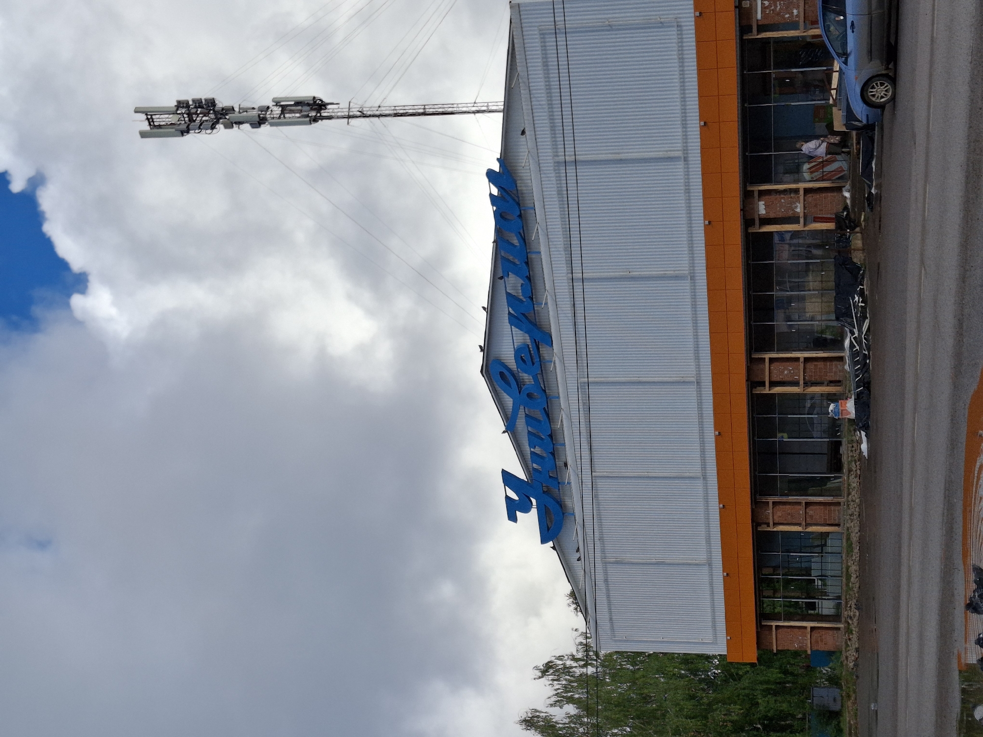 The image shows a building with a blue Cyrillic sign that reads "Универмаг" (Univermag), meaning "Department Store." The building has a corrugated metal facade on its upper section and an orange band above a row of large windows on the lower section. The windows appear to be under construction or renovation, with visible wooden framing and some debris. A cell tower with multiple antennas is visible in the background, against a cloudy sky with patches of blue. Trees are present to the left of the building. A blue car is parked on the right side of the image, and a person is standing near the entrance of the building. The foreground shows a wet road with reflections and some dark debris. The scene is located in the city of Elovo, Russia.