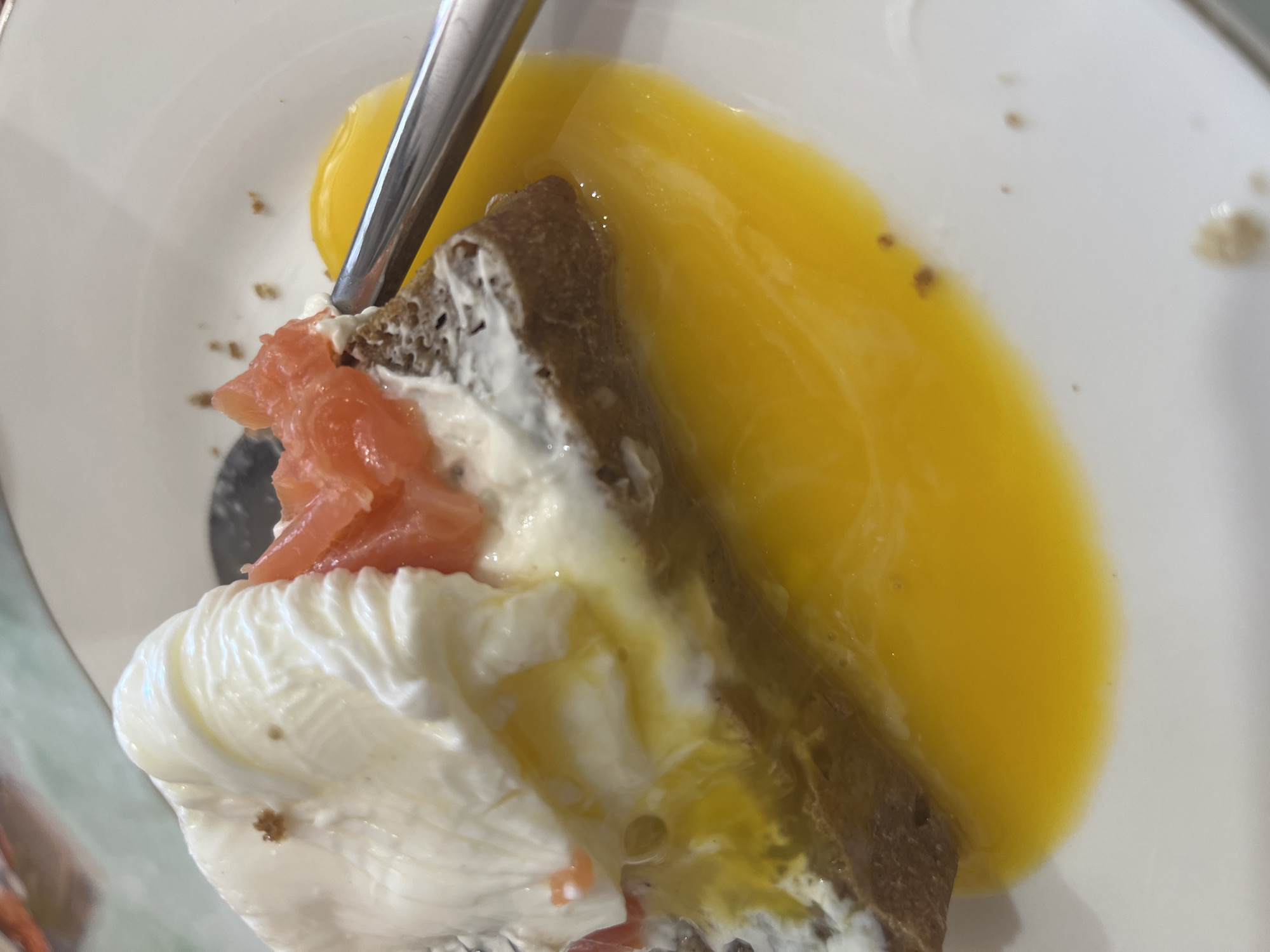 A close-up, overhead shot captures a serving of food on a white ceramic plate, likely depicting a breakfast or brunch item. The main subject is a piece of dark-colored bread or toast, topped with a generous layer of white, creamy spread, possibly cream cheese. Several pieces of thinly sliced, pinkish-orange smoked salmon are arranged on top of the cream.
A key element of the dish is a soft-cooked egg, likely poached, as a substantial amount of bright yellow, runny yolk has spilled onto the plate and is mingling with the other ingredients. A silver metal utensil, possibly a fork or spoon, is partially visible, inserted into the food, suggesting it is either being prepared for consumption or is in the process of being eaten.
The white plate shows some small brown specks, which could be seasoning like pepper, or crumbs from the bread. The overall presentation, despite the spilled yolk, suggests a rich and appetizing meal. There are no people visible, and no text is present in the image. The setting appears to be an indoor dining environment. The specific location of Chaikovsky, Russia, is provided as context, though no visual cues within the image directly confirm this.