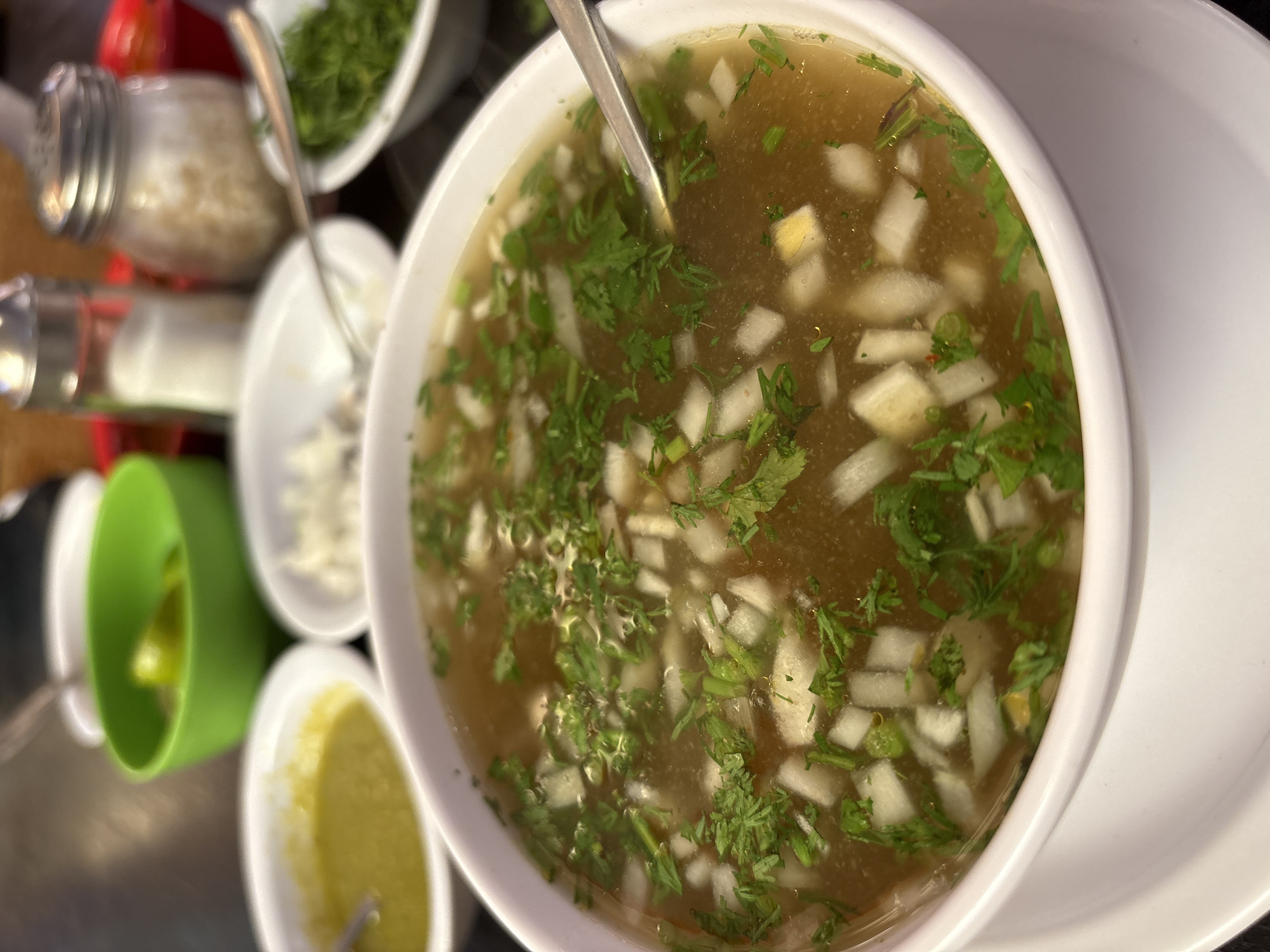 A close-up shot depicts a bowl of chicken soup at a restaurant in Ocotepec, Mexico. The bowl, which is white, contains a clear broth with diced white onions and chopped cilantro. A spoon is submerged in the soup. To the side, out-of-focus elements include condiment containers with green and white components, potentially containing additional garnishes. Salt and garlic shakers are also present in the image.