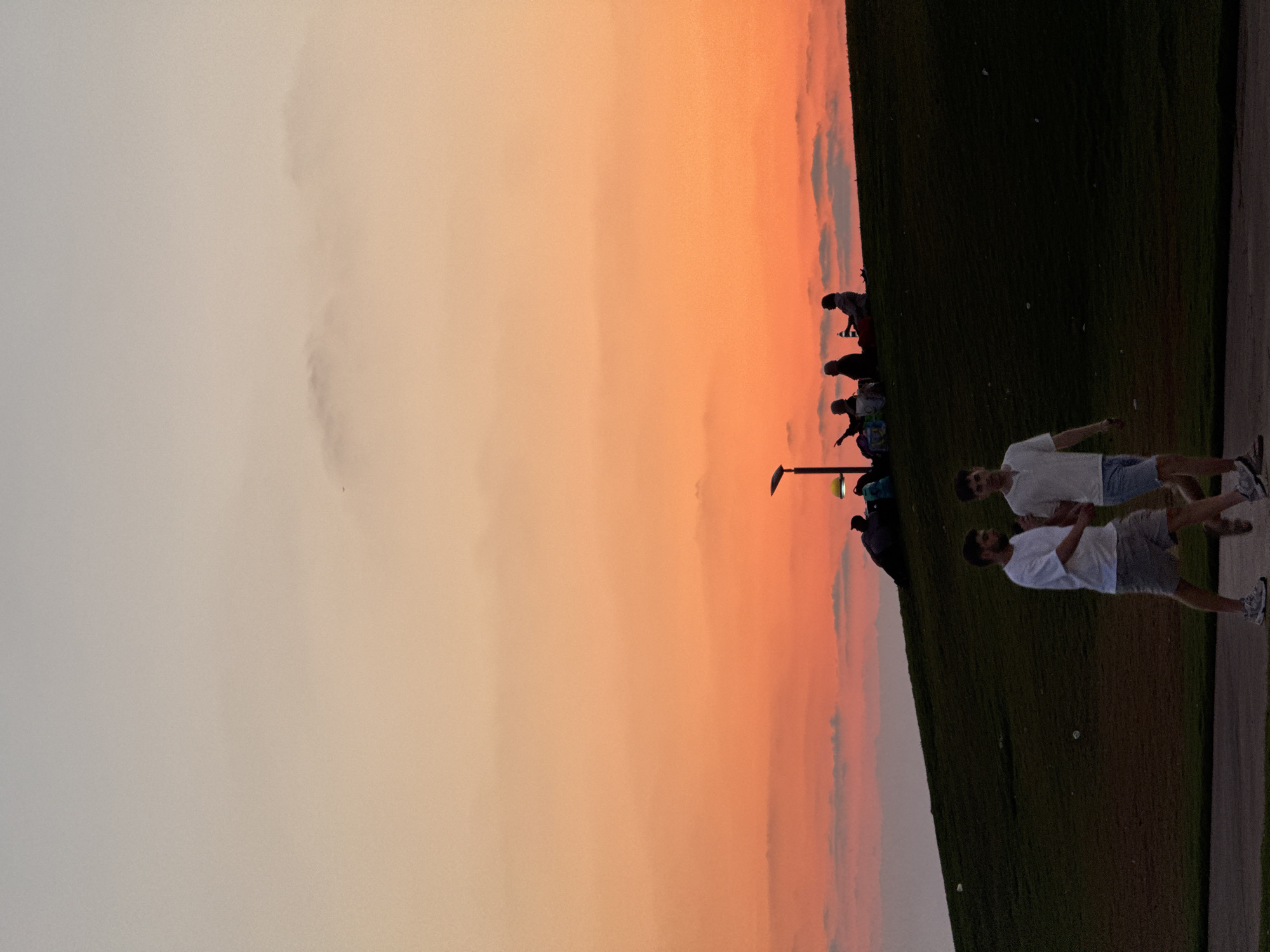 A low-angle, full-shot captures a sunset scene at a park in Tel Aviv, Israel. In the foreground, two young men walk towards the right side of the frame. The man on the left wears a white t-shirt and grey shorts, while the man on the right wears a white t-shirt and blue shorts. They are walking on a paved path with a grassy slope to their right. The slope is dotted with small white pieces of debris.
Further up the slope, silhouetted against the vibrant orange and pink sky, a group of people are gathered. Some are sitting on the grass, while others appear to be lying down. A lamppost with a yellow light fixture stands to the left of the group. The sky transitions from a pale orange at the horizon to a lighter, grayish hue at the top of the frame, with wisps of clouds scattered throughout.