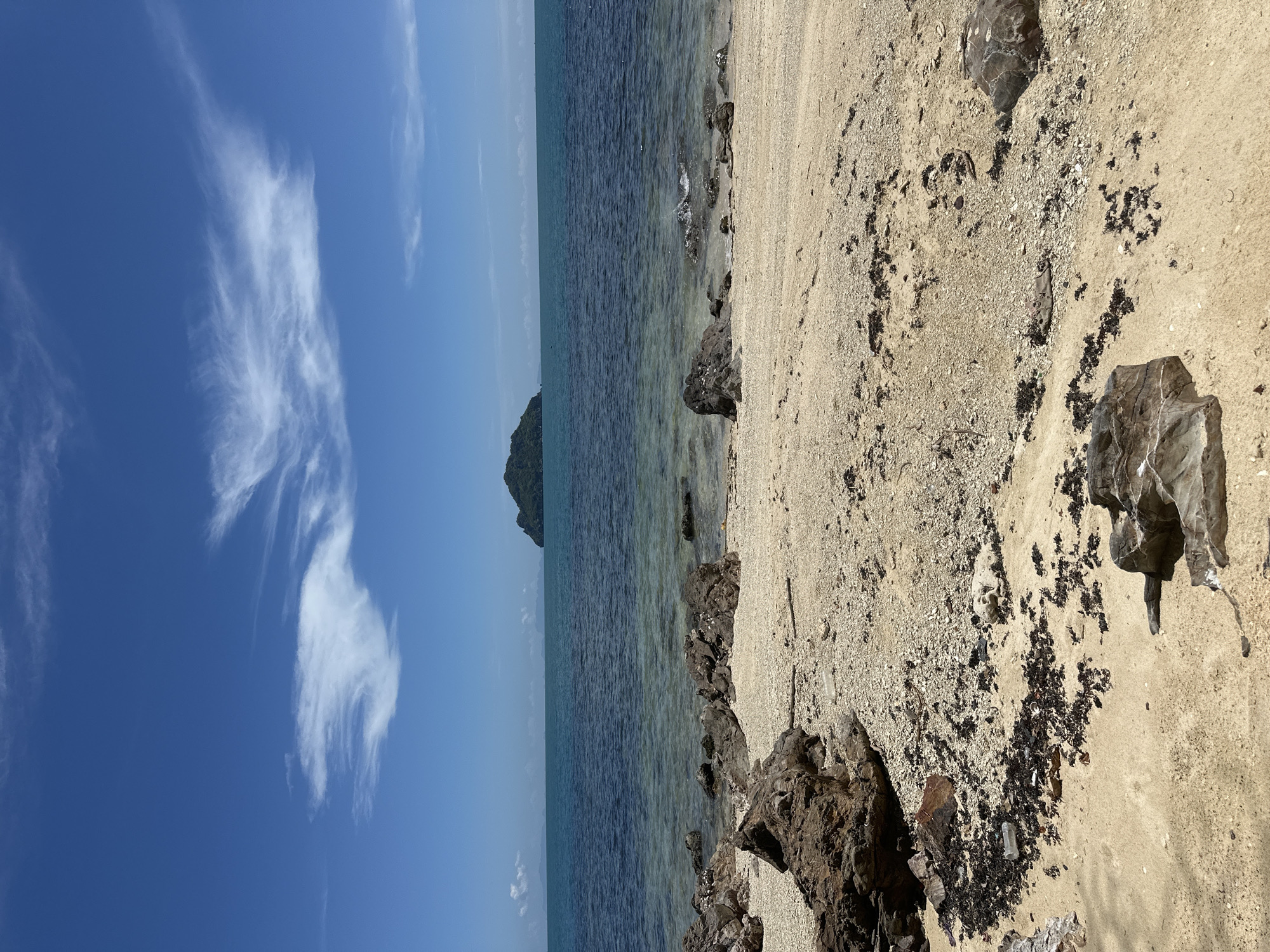 The image depicts a beach scene on Koh Phangan, Thailand. In the foreground, a sandy beach is strewn with rocks, seaweed, and some debris. The water is clear and turquoise, with small waves lapping at the shore. In the distance, a small, tree-covered island is visible on the horizon. The sky is a vibrant blue, with a few wispy white clouds. The overall impression is one of a tranquil and beautiful tropical paradise.