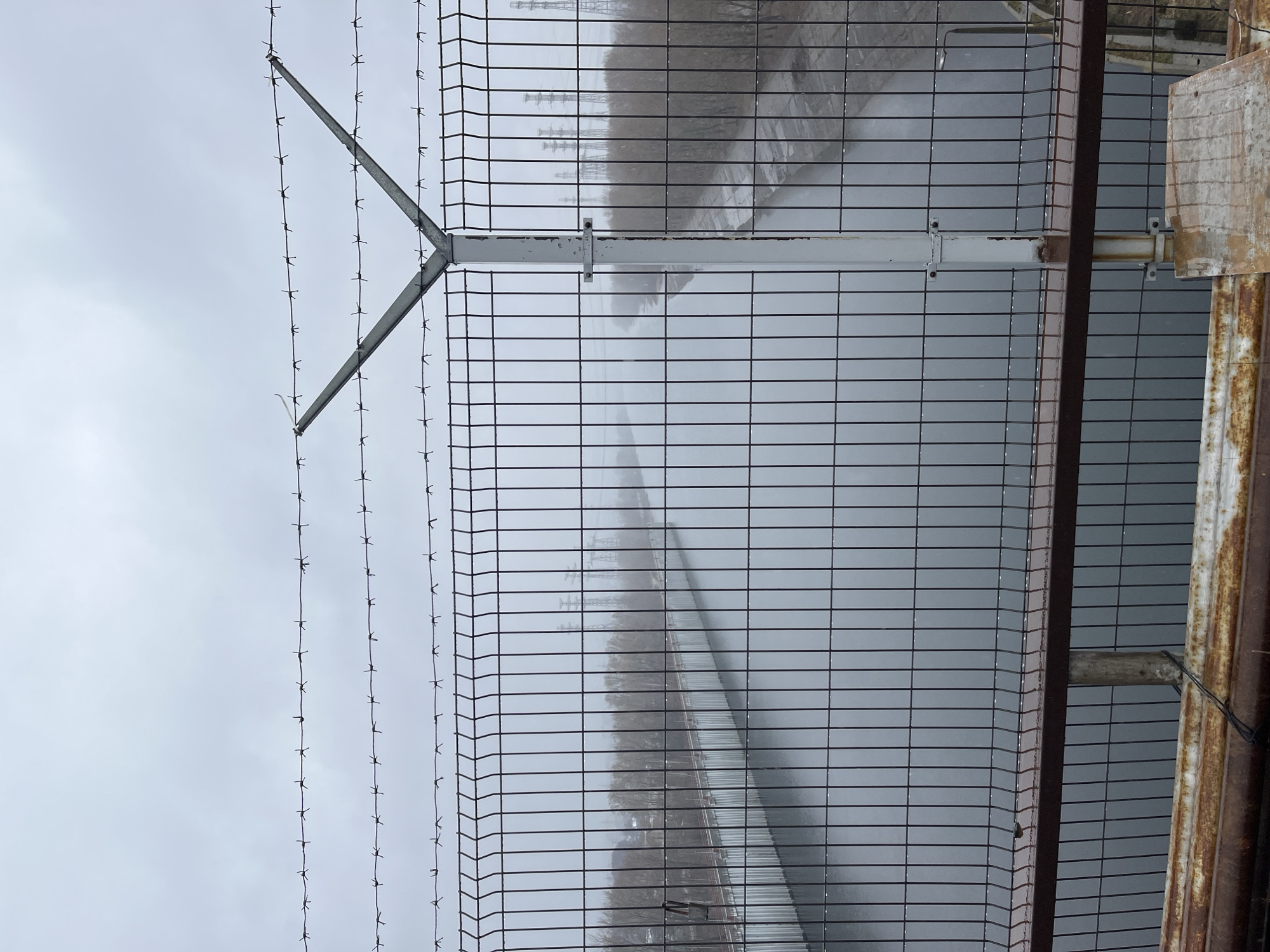 A prominent security fence, featuring dark metal mesh panels, dominates the foreground of this image. The fence is topped with multiple strands of barbed wire, supported by a 'Y'-shaped metal arm extending outwards from a white horizontal bar. This white bar runs horizontally across the fence panels, providing structural support and acting as a mounting point for the barbed wire arm. To the far right, a rusty, vertical metal post or column suggests another part of a larger structure or a continuation of the fence line.
Beyond the fence, a wide body of water, likely a river or reservoir, stretches into the hazy distance, its surface reflecting the dull sky. On the far bank of this water, a line of bare trees or vegetation is visible. Further in the background, several tall, dark industrial structures, resembling electricity pylons or parts of an industrial complex, rise faintly through the thick atmospheric haze.
The sky is uniformly overcast, contributing to a muted, low-contrast palette of grays and blues. The weather appears to be foggy or extremely hazy, severely reducing visibility in the distance and obscuring details of the landscape beyond the immediate vicinity of the fence. There is no discernible activity or event taking place; the scene is static and devoid of people. The overall impression is one of a somber, restricted industrial or infrastructural area, consistent with locations found in regions like Chaikovsky, Russia, which is known for its proximity to large waterways and industrial facilities. No visible text is present in the image.
