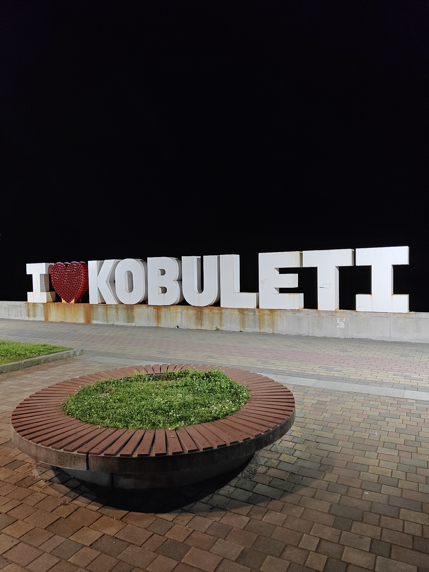At night, a ground-level shot shows the "I (heart) KOBULETI" sign in Kobuleti. The letters are large, white, and three-dimensional. A red heart symbol replaces the word "love." The sign sits on a short concrete wall or platform. Dark stains streak the wall behind the lettering. The background is black.
In the foreground, a circular wooden bench surrounds a raised flower bed. The bench has a concrete base with reddish-brown wooden slats forming the seating area. The flower bed in the center of the bench contains low-growing green plants with small white flowers. The ground is paved with multi-colored bricks.