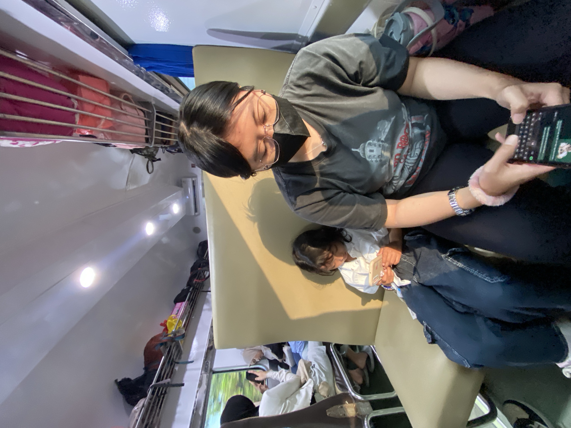Inside a train carriage, a woman wearing a black t-shirt, glasses, and a face mask is seated and looking at a phone. A young child in a white shirt is leaning against the woman. Passengers are visible in the background. Overhead luggage racks are present. The image appears to be taken during daytime. The location is Wonokoyo, Indonesia.