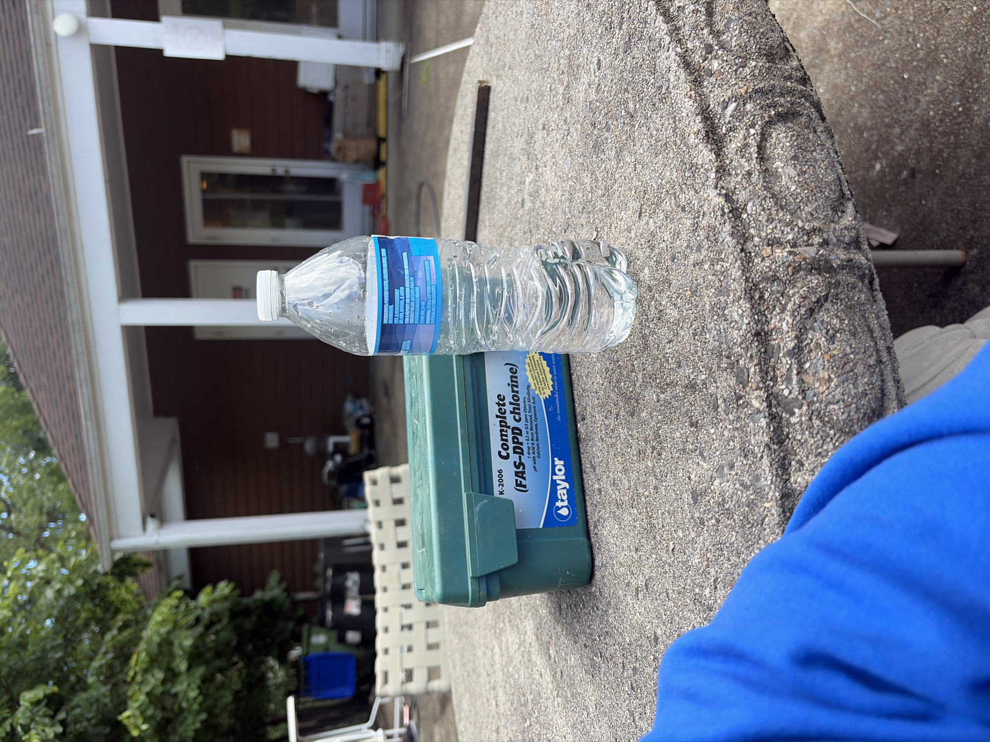A close-up shot shows a clear plastic water bottle resting horizontally on a concrete surface, next to a green "Taylor" pool water testing kit. The kit's label reads "K-2006 Complete (FAS-DPD chlorine)". A blue article of clothing is visible in the bottom right corner.
The scene appears to be outdoors, possibly a backyard or patio setting, with a building featuring white pillars and a brown exterior visible in the background. The presence of trees and lawn furniture further suggests an outdoor residential environment. It is daytime and sunny. Based on the OCR text "1 drop 0.2 or 0.5 ppm Chlorine, pH with Acid & Base Demand, Total Alkalinity, Calcium Hardness, Cyanuric Acid", the location could be in Edison, United States of America, a suburb of New York, New Jersey, where homes typically feature pools in their backyards.