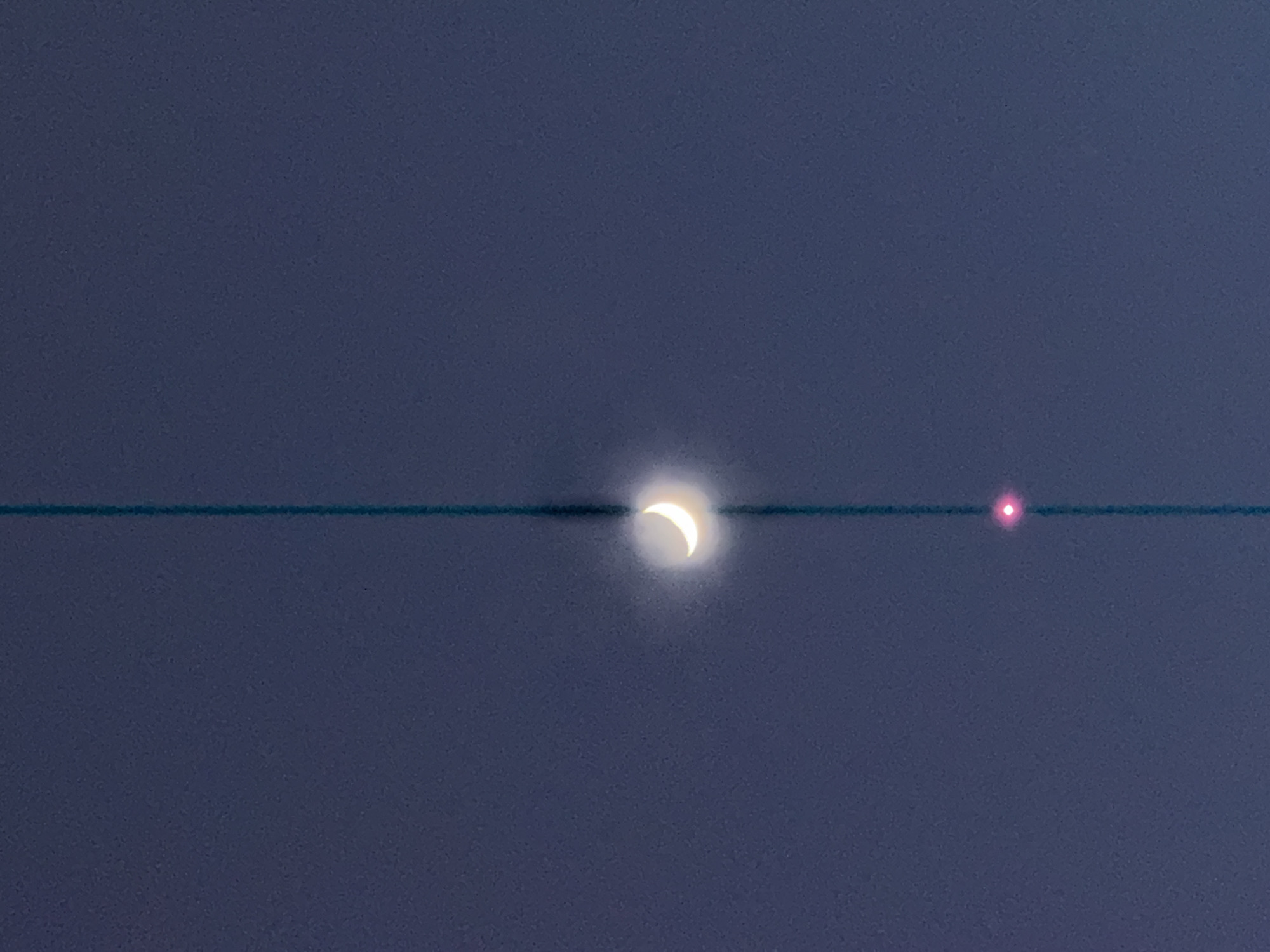 The image captures a celestial view against a dark blue-gray sky, suggesting twilight or nighttime conditions. The central subject is a bright, white crescent moon, exhibiting a soft glow or halo around its illuminated portion. To the right of the moon, a small, distinct, and intensely red light source or orb is visible.
A prominent, thin, dark blue-green horizontal line stretches across the entire width of the image, positioned directly in front of and bisecting both the moon and the red light. This line appears to be a physical obstruction, such as an overhead wire or cable, situated closer to the camera than the distant celestial objects. The way the line passes through the moon and the red light is a notable detail, creating a visual juxtaposition. The sky itself is clear, allowing for unobstructed viewing of the moon and the bright red light. No other objects, structures, or text are visible within the frame. The location context for this image is Khimki, Russia.