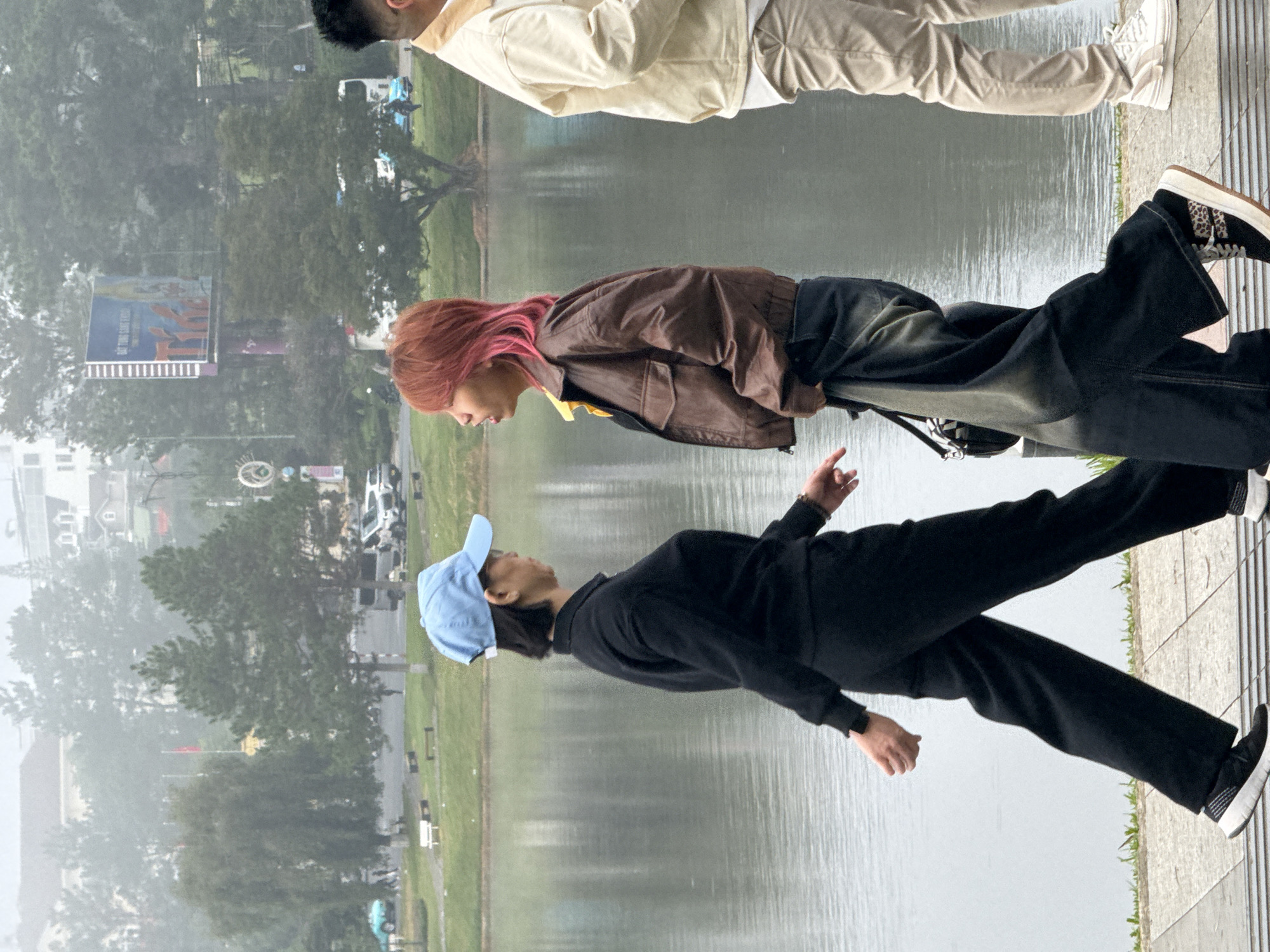 The image captures a scene along the edge of Xuan Huong Lake in Da Lat, Vietnam. Two individuals are walking on a paved pathway beside the lake. The person on the left is wearing a black outfit, including a long-sleeved top and wide-legged pants, and a blue baseball cap. They are mid-stride, with their left leg forward and their arms slightly bent. The person on the right is wearing a brown leather jacket over a yellow shirt and dark, baggy jeans, also walking. Their right leg is forward, and they are holding a black bag. Further to the right, another person is visible from the waist down, wearing a cream-colored jacket and light-colored pants, walking away from the viewer. The lake is calm with ripples reflecting the overcast sky and the surrounding greenery. In the background, trees and buildings are visible, partially obscured by a light mist, common in Da Lat's higher altitude. A billboard is visible in the distance above the trees. The atmosphere appears to be cool and possibly damp.