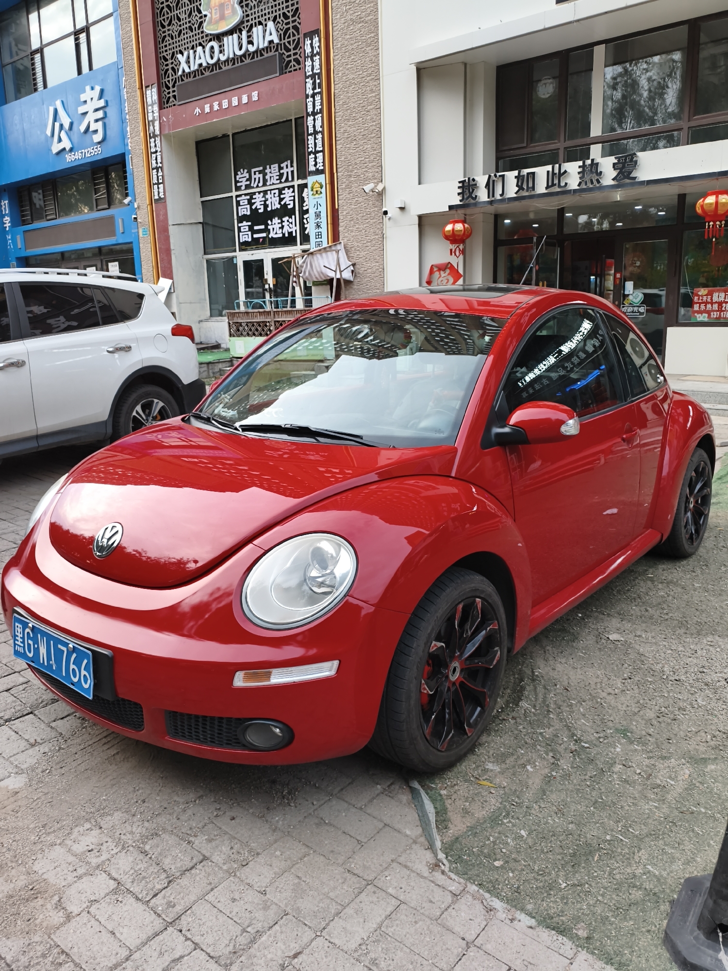 A bright red Volkswagen Beetle is parked on a sidewalk in front of a building. The car has a black and red rimmed wheel and a blue license plate with white lettering. The building behind the car has various signs with Chinese characters. The sidewalk is made of grey bricks. To the left of the Beetle, a white SUV is parked. The environment appears to be urban, possibly a commercial area.