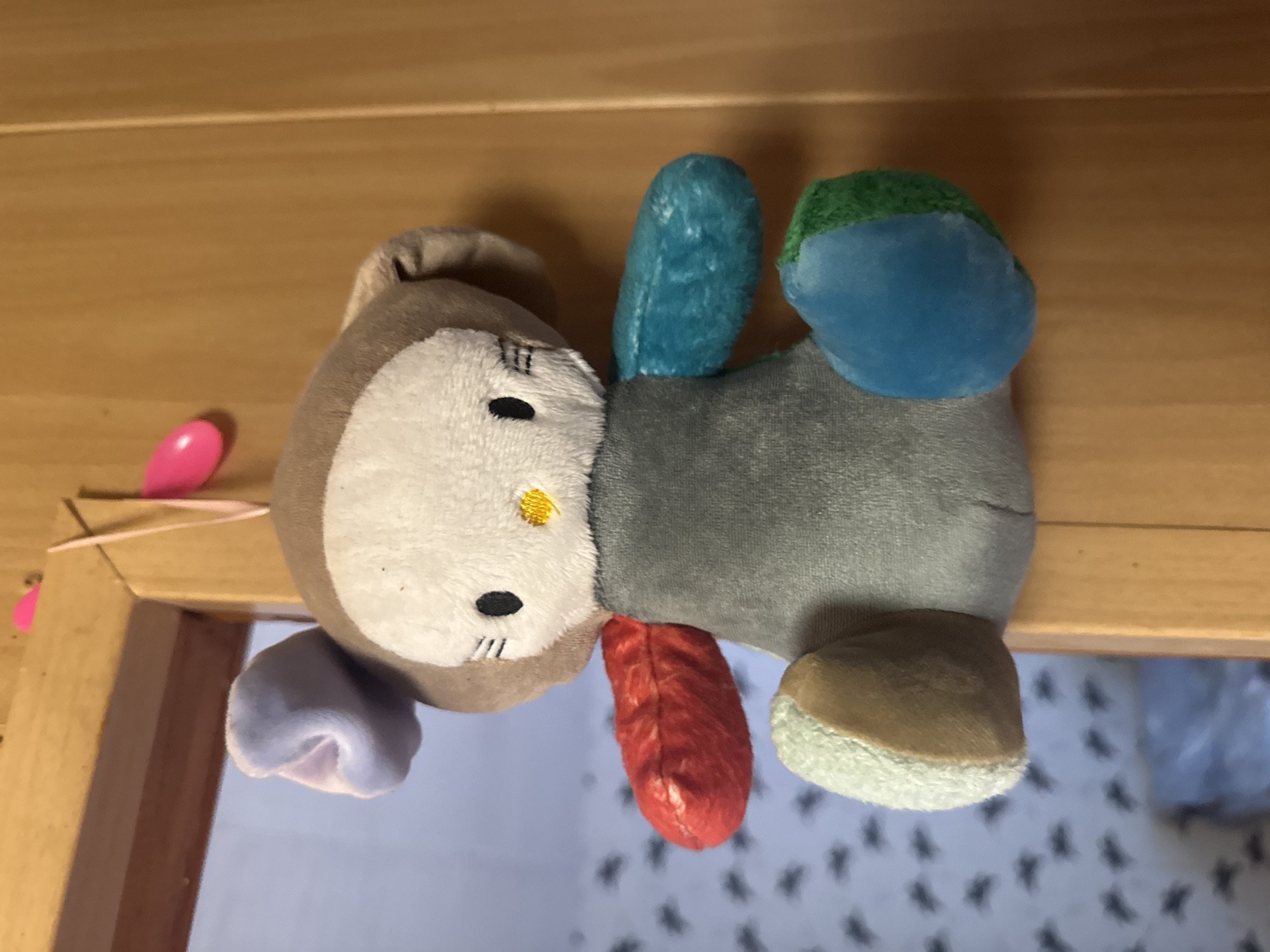 A close-up, eye-level shot shows a small, multi-colored plush toy hanging from a pink ribbon and pink plastic clip attached to a wooden doorframe. The toy resembles a character with a white, round face, black embroidered eyes and mouth, and a yellow embroidered nose. It has large, light purple ears. The body of the toy is a patchwork of different colored fabric: red on its left arm, grey on its torso, turquoise on its right arm, light green on its left leg, and blue and green on its right leg. The background on the left is a blurry wall with a repeating pattern of small, dark figures. The background on the right is a wooden door. The image is lit with a bright light from the front. This scene is located in Shcherbinka, Russia.