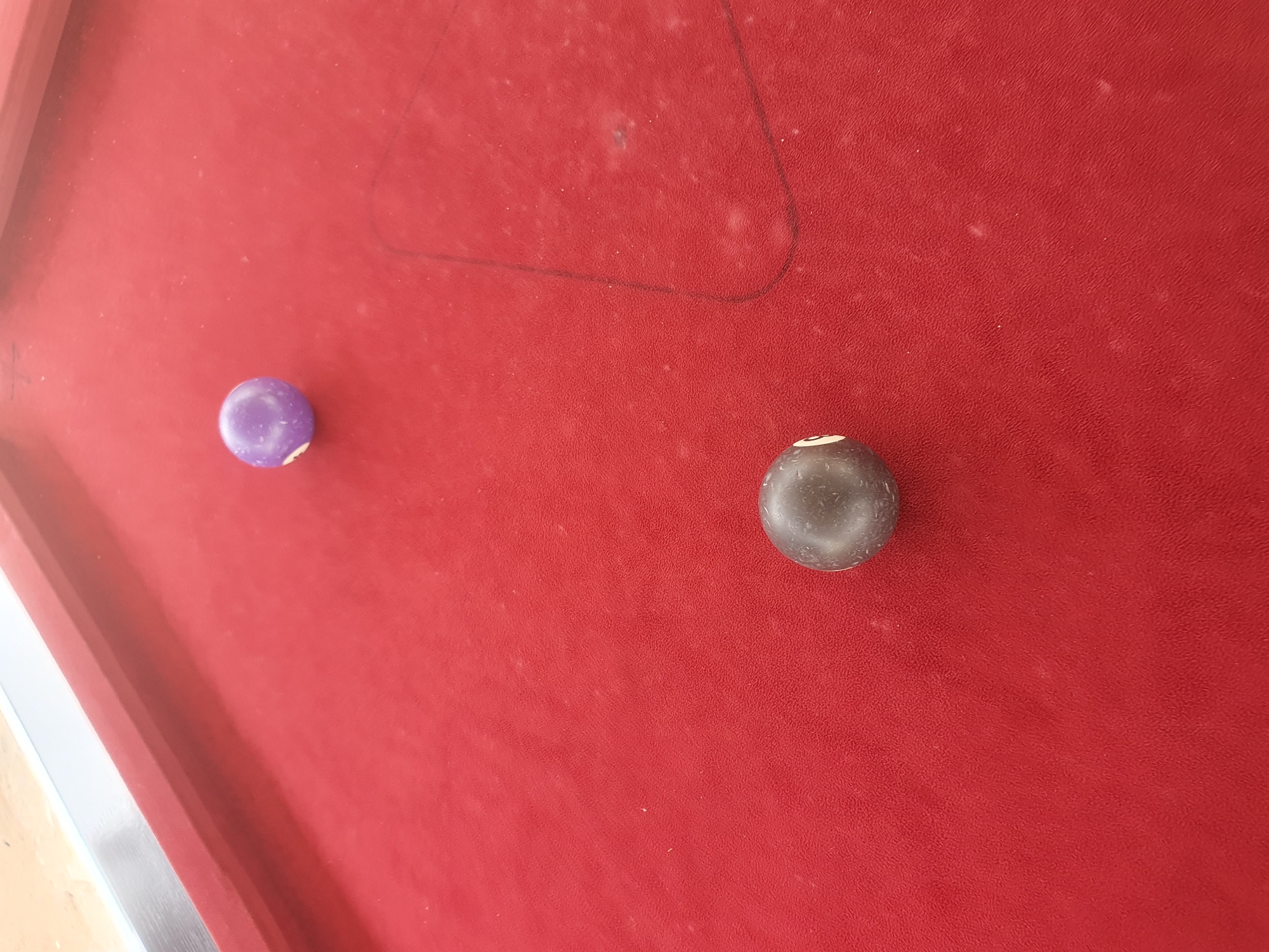 An overhead shot of a red billiard table with two balls. One ball is purple with a white number 9 on it, and the other ball is black with a white number 8 on it. The table is dusty and shows a faint triangle outline of where the balls would typically be racked. The scene is set in Lokoja, Nigeria.