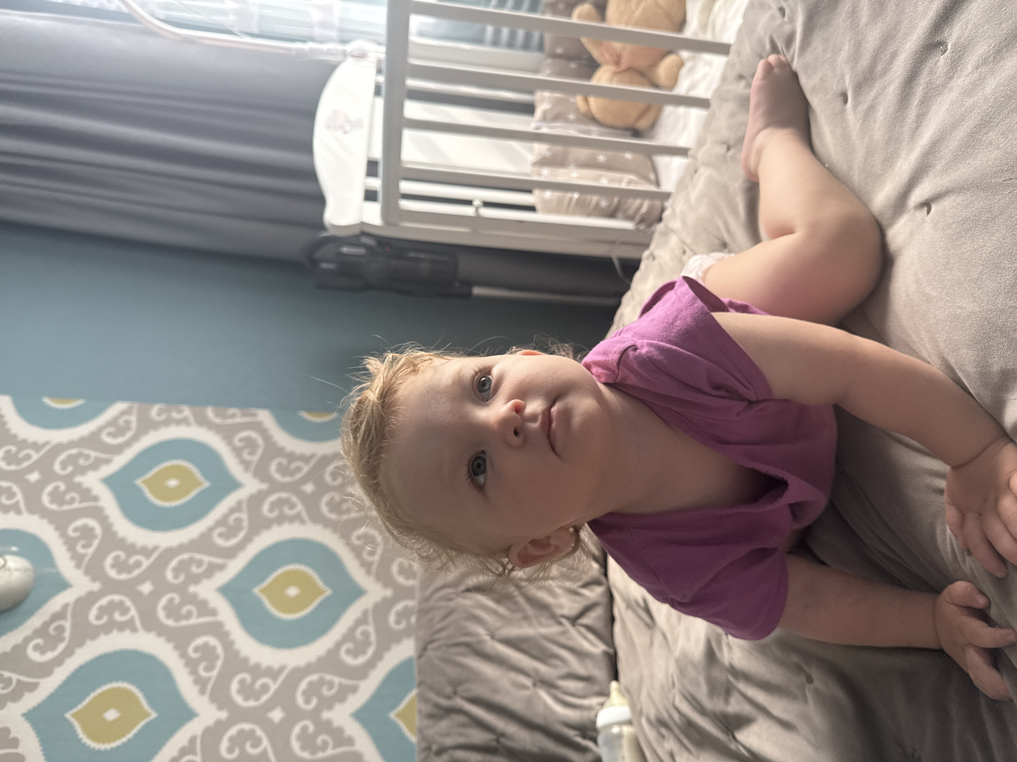 A Caucasian toddler is on a bed, on their hands and knees, wearing a purple t-shirt and diaper. The toddler has blonde curly hair and blue eyes, and is looking to the right of the frame. Behind the toddler, there is a grey tufted duvet, a pillow, a baby bottle, and a white crib with a teddy bear and pillows inside. The wall behind the bed has a pattern of grey, white, and yellow shapes. The room is lit by natural light coming from a window to the right. The location of Kondopoga, Russia is not discernable from the image.
