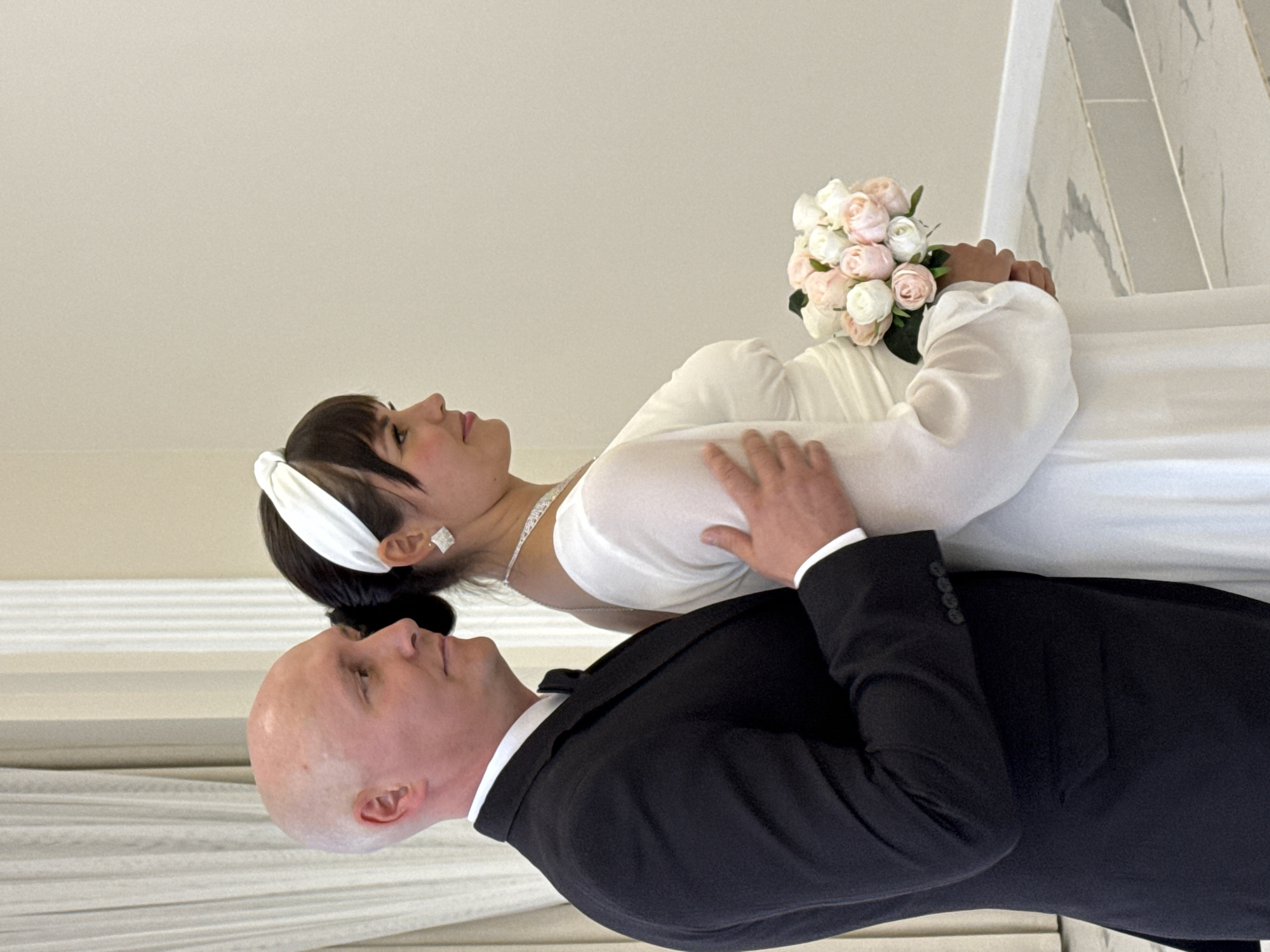 A medium shot shows a bald man in a black tuxedo and a woman in a white wedding dress, both facing right, on what appears to be a wedding day. The man's left arm is around the woman's waist. The woman holds a bouquet of pink and white roses in her right hand. The background is a plain, light-colored wall with pleated drapes to the left. The floor is a light gray, marble-patterned surface with steps. The scene appears to be indoors in a formal setting, possibly a wedding venue.