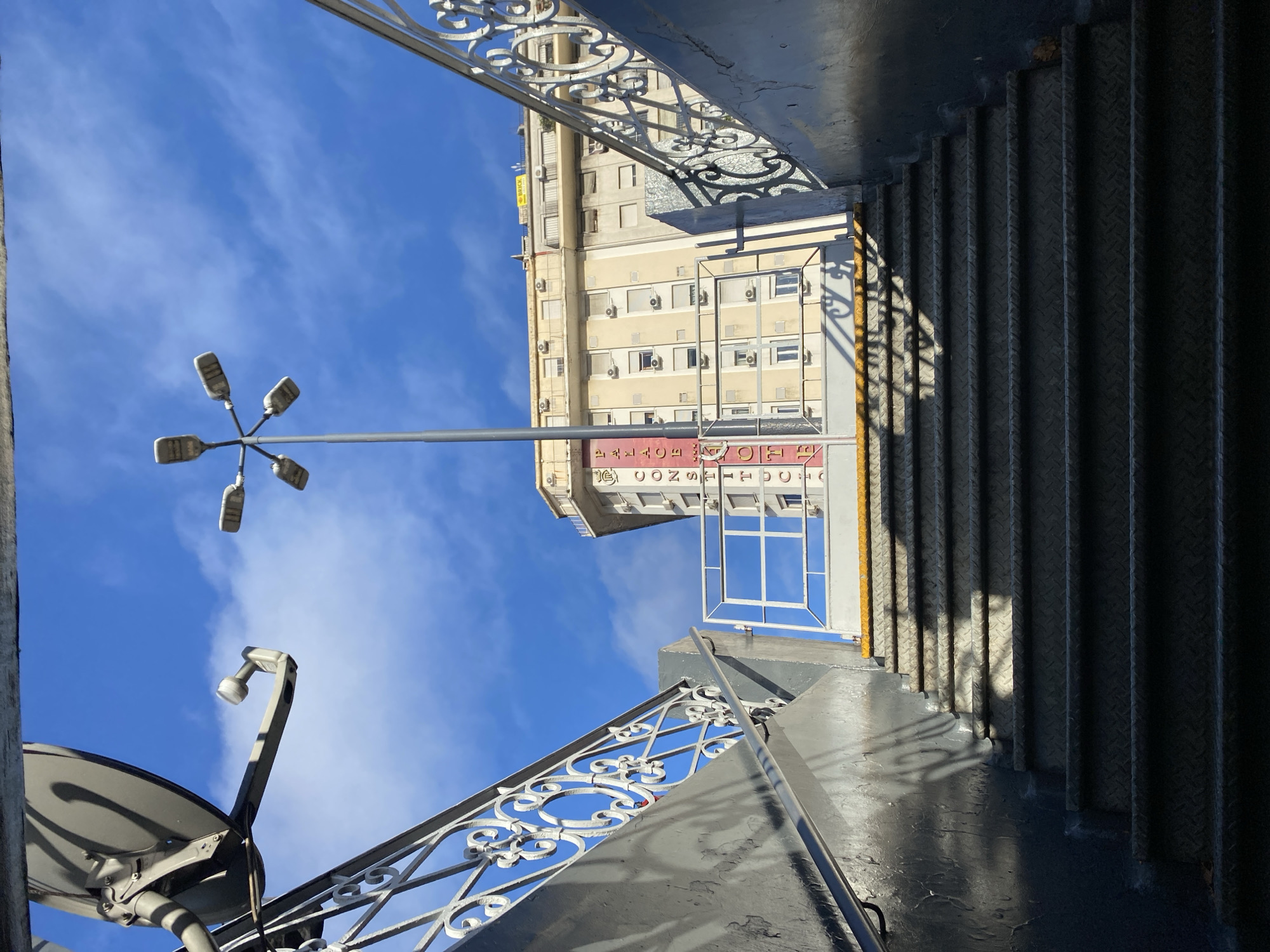 The image presents a dramatic, low-angle perspective looking sharply upwards towards a clear blue sky in an urban setting, likely Buenos Aires, Argentina.
On the right side of the frame, a dark, metallic staircase with textured diamond plate steps descends, partially obscured by deep shadow. A decorative, white metal railing with ornate scrollwork runs along the edge of the staircase, casting intricate shadows onto the steps and a lower platform, indicating strong, direct sunlight.
Against the expansive blue sky, a tall, slender street lamp post with four multi-directional luminaires is prominently featured on the left. Further down and to the left, a curved, gray structure, possibly part of another light fixture or a small satellite dish, is partially visible.
In the middle and upper-left, a multi-story building with a light yellow-beige facade is visible. It features numerous windows and several outdoor air conditioning units. Below the building's main structure, a red sign is partially discernible. When the image is mentally rotated, the text appears to read "HOTEL CONSTITUCIÓN." Higher up on the building, a smaller yellow sign with black lettering reads "BRICK."
The background is dominated by a vibrant blue sky, dotted with sparse, wispy white clouds. The strong sunlight creates clear contrasts and pronounced shadows, suggesting it is daytime. The overall impression is one of architectural detail and urban infrastructure viewed from an unusual, upward-gazing angle.