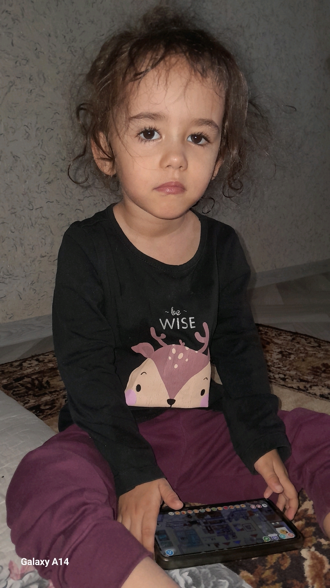 In Karshi City, Uzbekistan, a young child with curly brown hair and a neutral expression sits on a carpeted surface, looking directly at the camera. The child is wearing a long-sleeved black shirt with a graphic of a deer and the text "~be wise~" and burgundy pants. The child's hands rest on a tablet device displaying a colorful interface, which is placed on their lap. The background consists of a textured wall and floor, with a patterned rug partially visible. The image is a medium shot, capturing the child from the chest up and the tablet in focus.