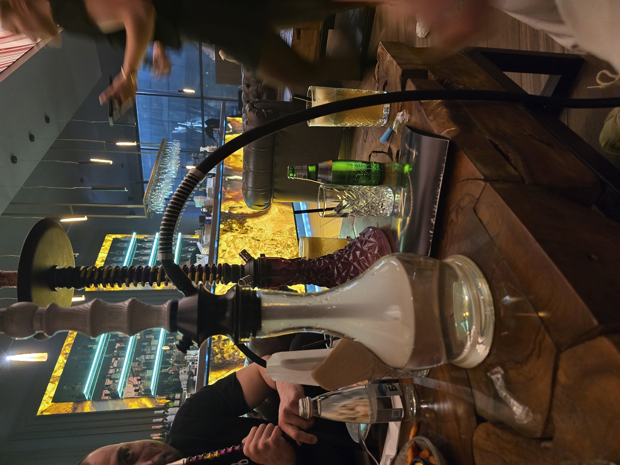 The image depicts an indoor scene, likely within a bar or lounge in Burgas, Bulgaria. Foreground features a hookah with a clear glass base filled with a milky substance, a black hose, and a decorative stem. A person in a black shirt is visible near the base, appearing to be preparing or using the hookah. The table on which the hookah rests is made of dark wood. In the background, a bar with illuminated shelves displaying bottles of alcohol and glasses is seen. Additional glassware, a green bottle of liquid, and a glass of amber-colored liquid are also on the table. People are present in the background, though their activities are not clear. The environment is dimly lit, creating a relaxed atmosphere.