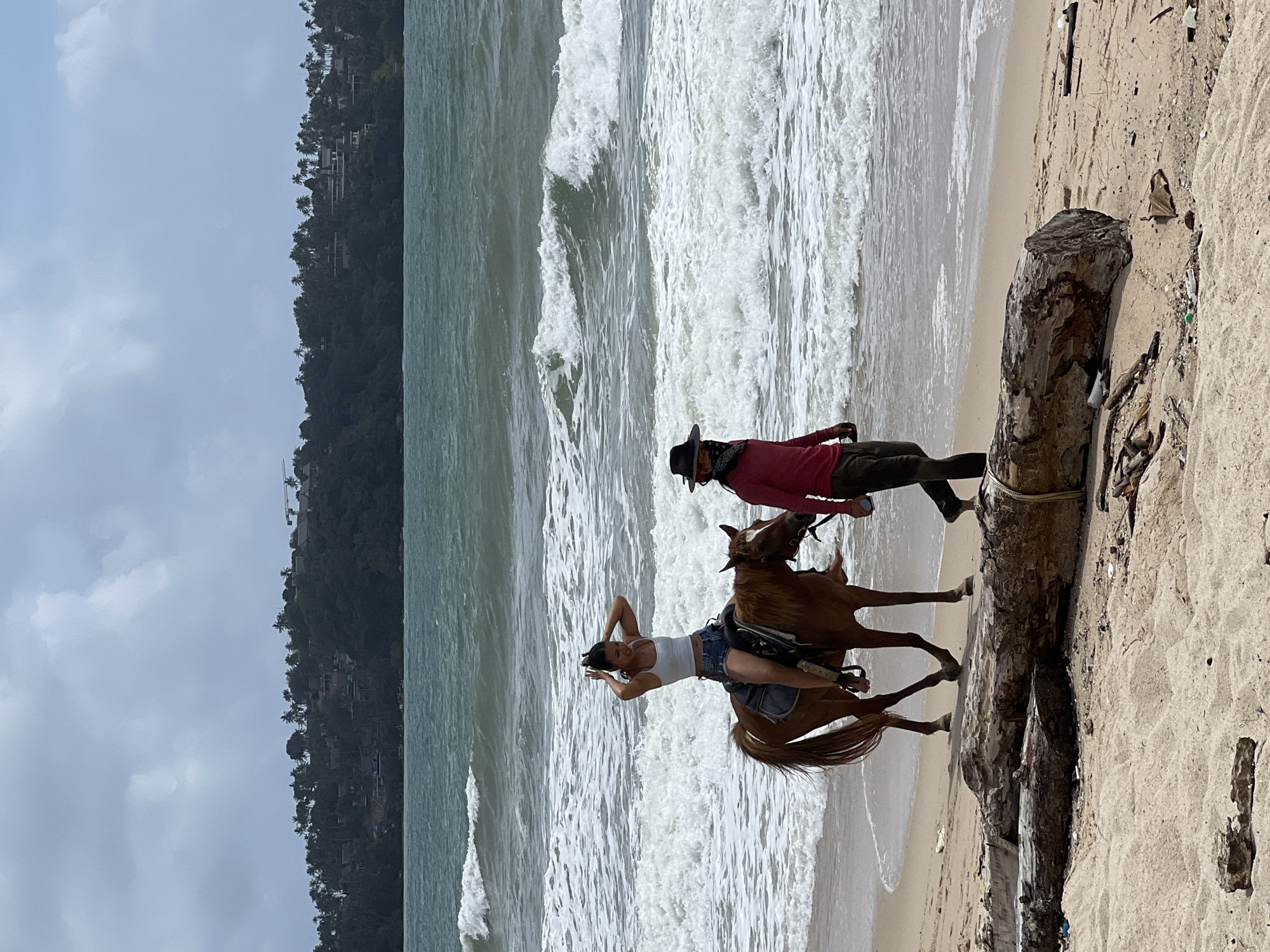 A woman on horseback and a man walking beside her are on a sandy beach. The woman is wearing a white tank top and denim shorts, and she is holding her hands to her hair. The horse is brown and appears to be saddled. The man is wearing a red long-sleeved shirt, dark pants, a cowboy hat, and a bandana over his face. They are walking along the shoreline, with waves from the ocean crashing behind them. In the background, a tree-covered hill with houses and a construction crane is visible under a cloudy sky. The foreground of the beach has a large piece of driftwood and scattered debris. The location is a beach in Choeng Thale, Thailand.