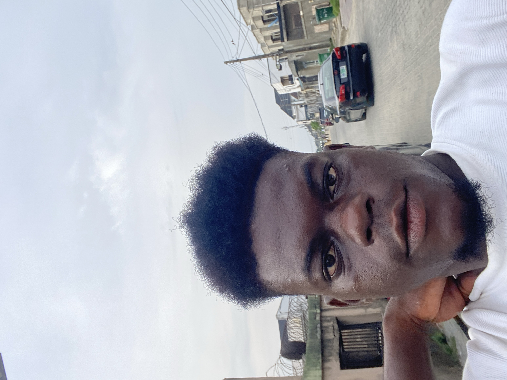 A close-up selfie of a dark-skinned man with short, curly black hair, taken outdoors during the daytime. The man is wearing a white ribbed shirt and is positioned at an angle in the frame, with his hand near his chin. The background includes a street scene with buildings, utility poles with wires, and parked cars. The sky is overcast. The location is in a city, though the specific city is unknown.