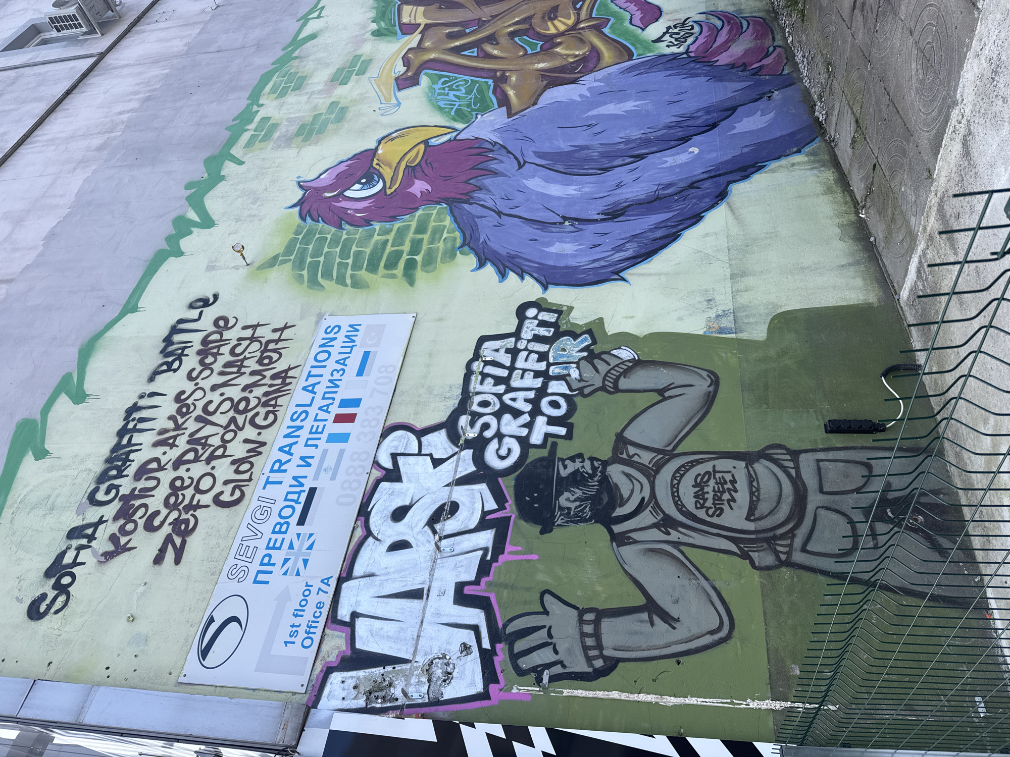 This is an outdoor scene depicting a wall covered in graffiti, likely located in Sofia, Bulgaria, as indicated by the "Sofia Graffiti Tour" lettering. The graffiti includes a large purple bird-like creature, stylized text, and a cartoonish figure of a man wearing a hat and vest with "RAYS STREET 222" written on it. A sign for "SEVGI TRANSLATIONS" is attached to the wall. A green fence runs along the bottom right of the frame. The scene is shot from a slightly tilted angle.