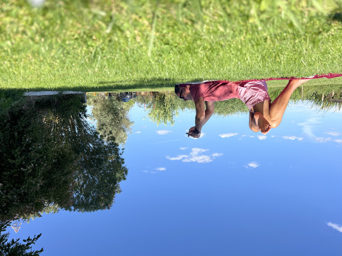 An adult male, wearing a red t-shirt and light pink shorts, lies supine on a red mat positioned on vibrant green grass. His left leg is bent with the foot flat on the mat, while his right ankle rests across his left thigh. Both arms are raised overhead, hands grasping a dark, rectangular object, possibly a mobile phone. He is barefoot and appears to be looking upwards at the object. The scene is outdoors under a clear blue sky with scattered white clouds, illuminated by direct sunlight, casting shadows. In the mid-ground, a line of dense green trees and bushes flanks the right side. A dark-colored vehicle, appearing to be an SUV, is partially visible in the distance, positioned on an unpaved path or driveway. To the left, tall green vegetation, possibly reeds or grasses, extends towards the horizon, with faint wooden posts visible. This scene is located in a rural or semi-rural area within Coruche, Portugal.