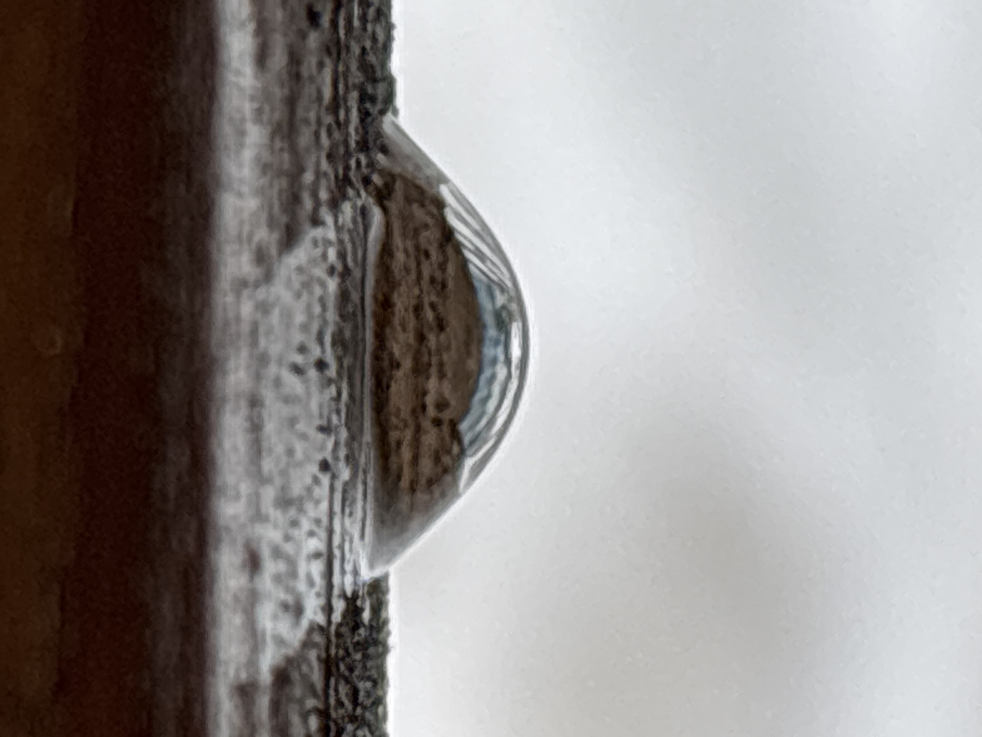 A close-up photograph shows a water droplet clinging to the edge of a dark, wooden structure, likely a window frame or post. The droplet distorts the view of what's behind it, suggesting a wet, overcast day. The background is a blurred, light gray, possibly indicating a neutral or pale-colored surface or an overcast sky. No people are visible in the image. The location is suggested to be Chaikovsky, Russia.