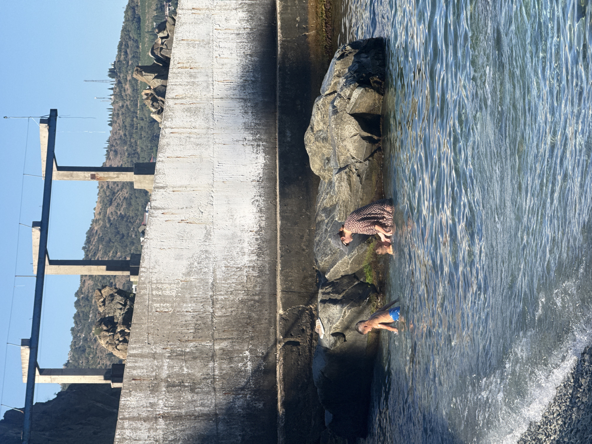 In Partenit, Ukraine, on a sunny day, a woman wearing a patterned dress and a cap is in the shallow waters of the sea, tending to two children. One child, wearing blue swim trunks, stands in the water. The second child, partially submerged, is near the woman. They are situated near large rocks on a shoreline adjacent to a weathered concrete structure. The sea is clear and calm, with small waves lapping at the pebble beach. In the background, a hilly landscape covered with sparse trees rises under a clear blue sky. The scene depicts a family enjoying a day at the beach near the iconic concrete piers.