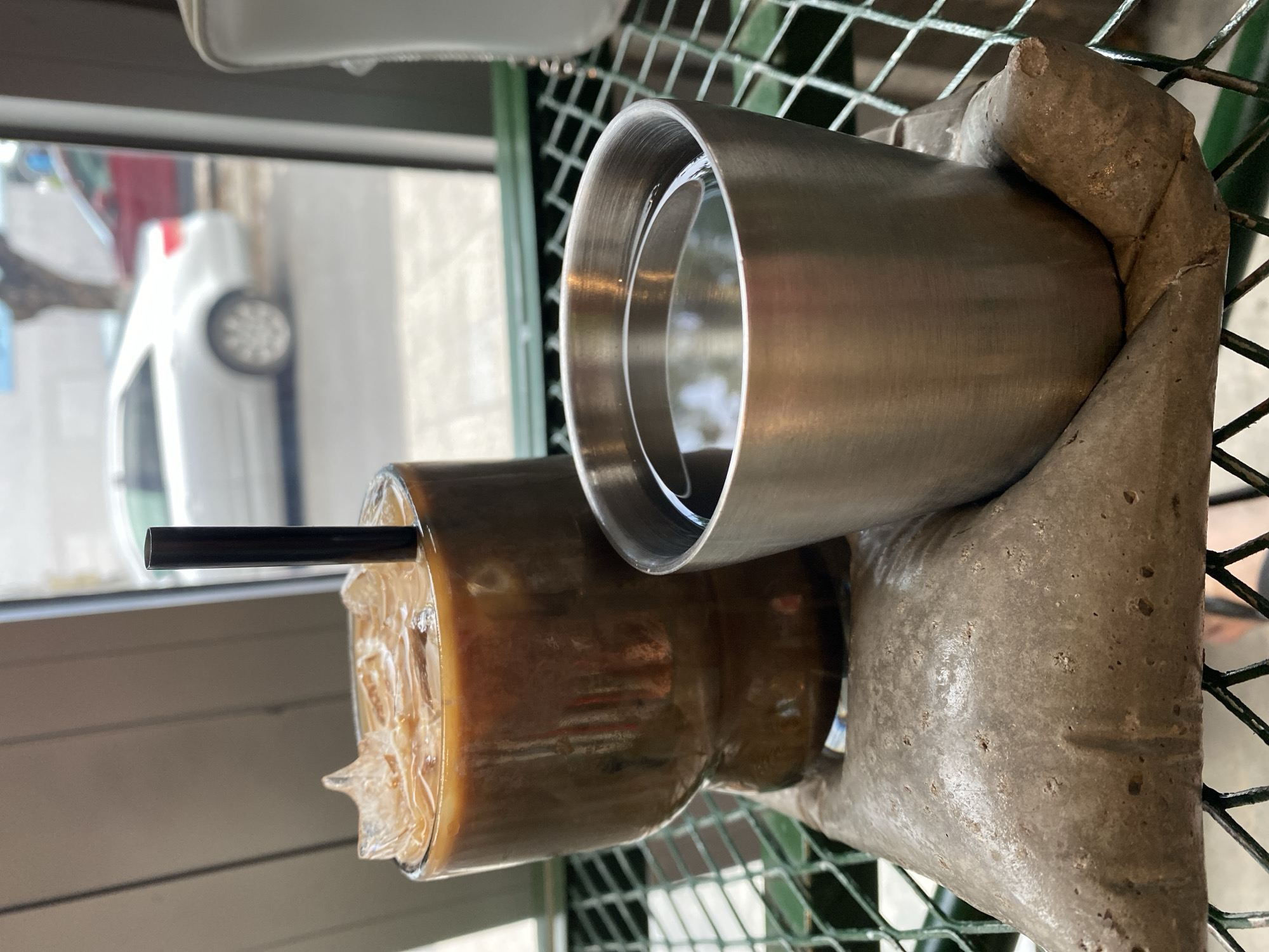 Two beverages, one in a clear glass with ice and a dark liquid, and another in a small metallic cup with a dark liquid, are placed on a textured grey object on a green metal grate table. Outside a window, a silver sedan and a red car are visible on a street. The scene appears to be in a cafe or restaurant in Da Nang, Vietnam, suggested by the presence of a Vietnamese-style sugar packet that is holding the drinks in place.