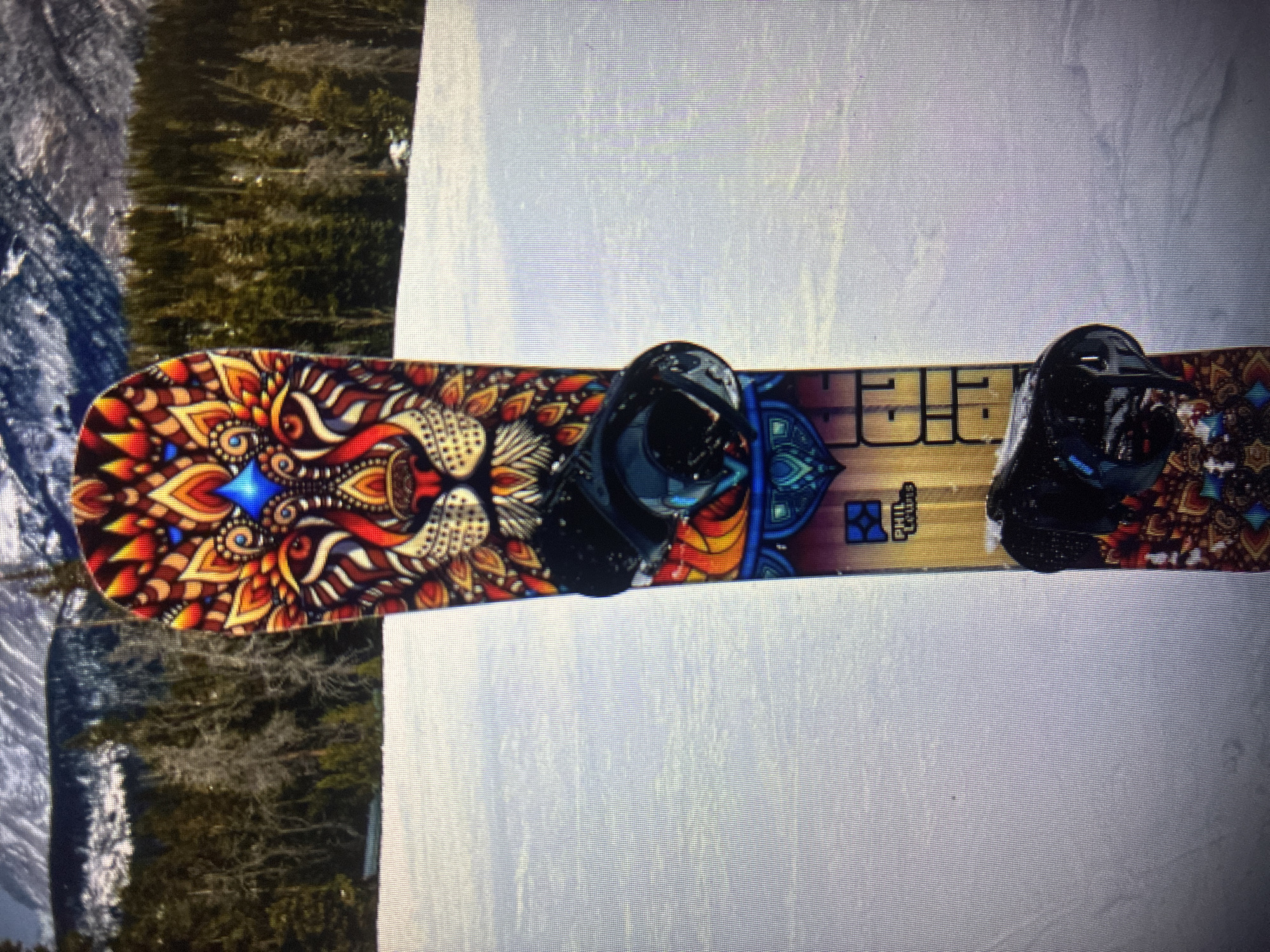 The image captures a vibrant snowboard resting on a snow-covered surface in a mountainous landscape. The snowboard is positioned horizontally across the frame. Its top features an elaborate, colorful design resembling a mandala, with a prominent, stylized lion's head or face detailed with patterns and jewel-like blue accents towards the nose (left side). The color palette is rich, with dominant reds, oranges, and yellows contrasting with blues and turquoise. Two black snowboard bindings are mounted on the board. Near the rear binding (right side), the brand name "STEP CHILD" is visible in a stylized font, alongside a small blue square graphic.
The setting is a bright, daytime winter scene. The snowboard lies on a smooth, white expanse of snow. In the blurred background, a dense forest of evergreen trees, possibly dusted with snow, extends up a mountain slope. The overall scene suggests a ski resort or a snowy trail suitable for snowboarding. No people are visible. This winter sports setting is consistent with the provided location context of Asenovgrad, Bulgaria.