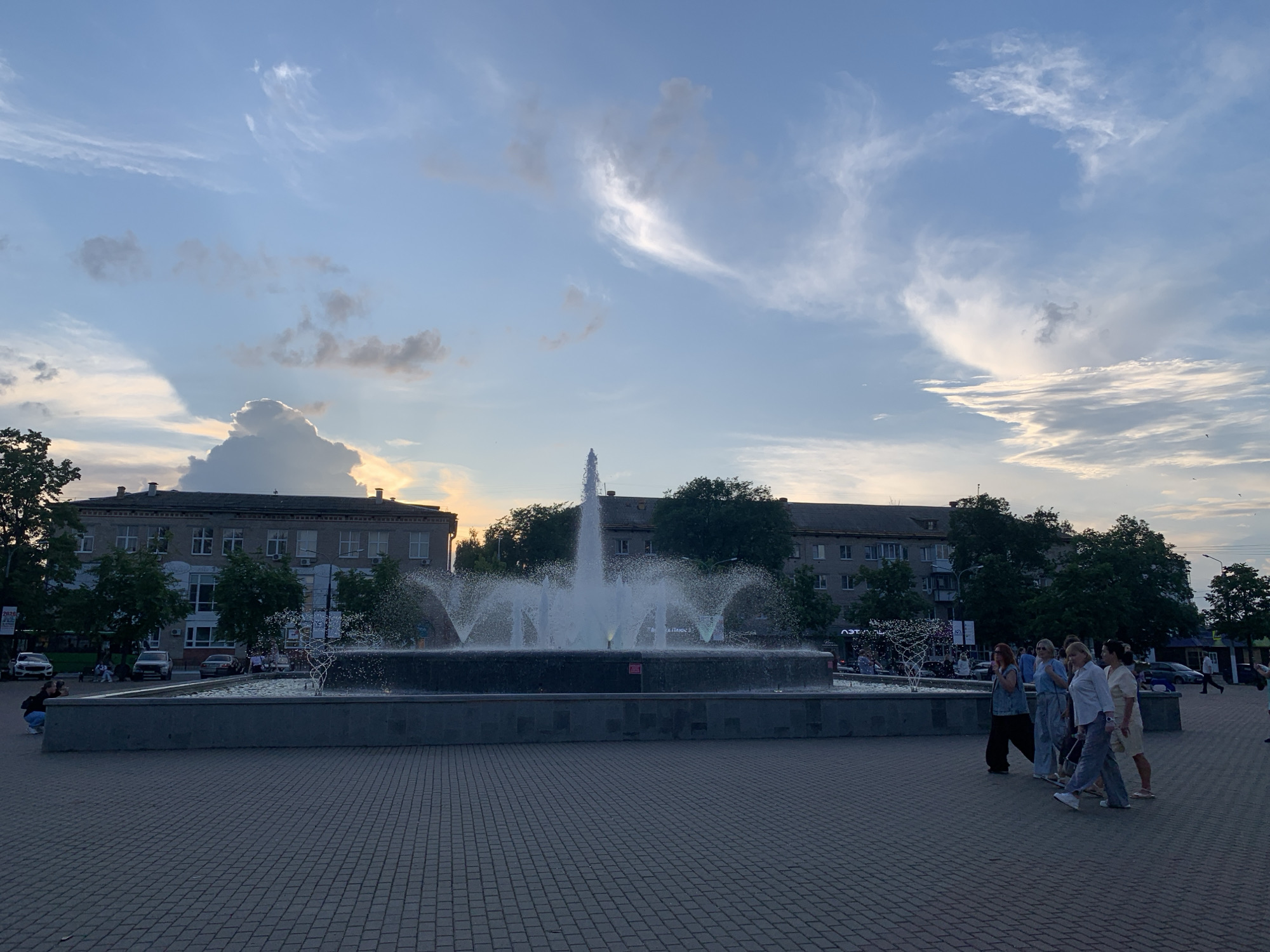 The image depicts a public square in Chaikovsky, Russia, featuring a large, multi-tiered fountain as its central element. The fountain is actively spraying water, creating a dynamic display against a backdrop of buildings and trees. Several individuals are visible in the foreground and midground, appearing to be walking or observing the fountain. Some are dressed in casual attire. The sky above is partly cloudy, with a soft, diffused light suggesting either early morning or late afternoon. The ground is paved with interlocking bricks. Buildings lining the square appear to be apartment complexes or commercial establishments. The overall scene conveys a sense of urban public space during a calm period.