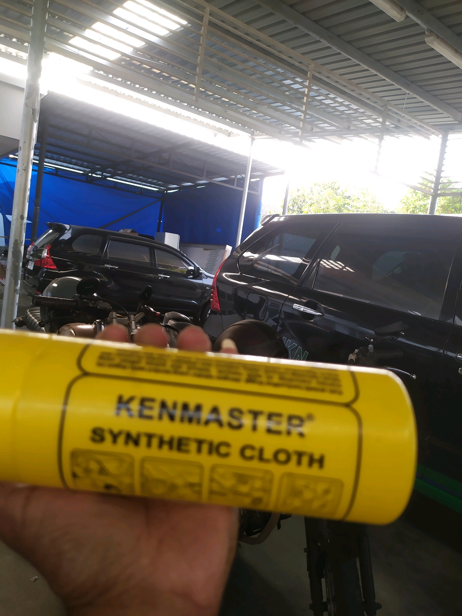 A person's hand is holding a yellow cylindrical object with text that reads "KENMASTER SYNTHETIC CLOTH". The background features two dark-colored vehicles, possibly cars or SUVs, parked under a roofed structure with a corrugated metal roof and fluorescent lighting. A blue tarp is visible behind the vehicles. The overall environment appears to be an outdoor or semi-outdoor workshop or parking area. The lighting suggests it is daytime with bright sunlight filtering through the roof.