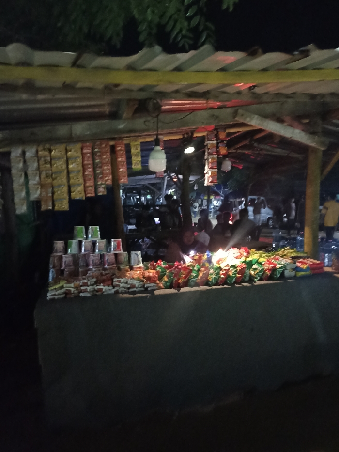 The image depicts a lively, open-air snack stall operating at night in Kecamatan Pujut, Indonesia. The stall is constructed informally with a corrugated metal roof, supported by a wooden or bamboo framework, and features a prominent yellow beam running along its front.
Artificial lighting, including a visible white bulb hanging from the roof, illuminates the stall and its offerings against the dark background of the night. Numerous individually packaged items, likely sachets of instant food or drinks, hang from the overhead structure. The main counter, draped in a dark fabric, displays a variety of products. On the left, stacks of disposable cups, some green and others with colorful patterns, are neatly arranged. The rest of the counter is covered with an abundance of packaged snacks like chips and biscuits, alongside bottles of what appear to be beverages on the far right.
Several people are visible in the scene, both behind the counter and in the background. One individual is more clearly discernible behind the counter, partially lit, seemingly attending to the stall's operations or engaging with customers. Other blurry figures and vehicles in the distance suggest a bustling roadside or market environment.
The time of day is distinctly night, creating a warm contrast between the well-lit stall and its dark surroundings. The atmosphere appears casual and active. No discernible text is visible on any of the products or stall signage.