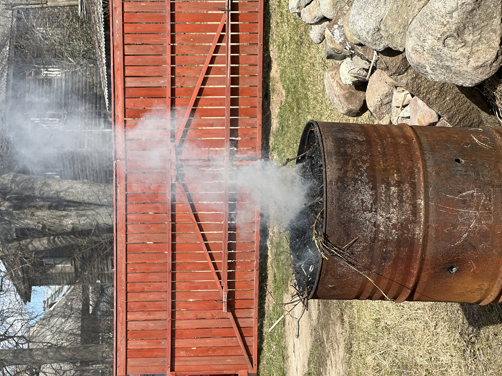 The image depicts a metal burn barrel positioned next to a red wooden fence. Smoke rises from the barrel, indicating that something is burning inside. To the right of the barrel, there is a pile of rocks. In the background, there is a building with a complex structure, potentially a house. The environment consists of grass and what appears to be early spring vegetation, suggesting a rural or suburban setting. While it is claimed that this image is from Чернышово, Russia, there is no definitive evidence in the image to confirm this.