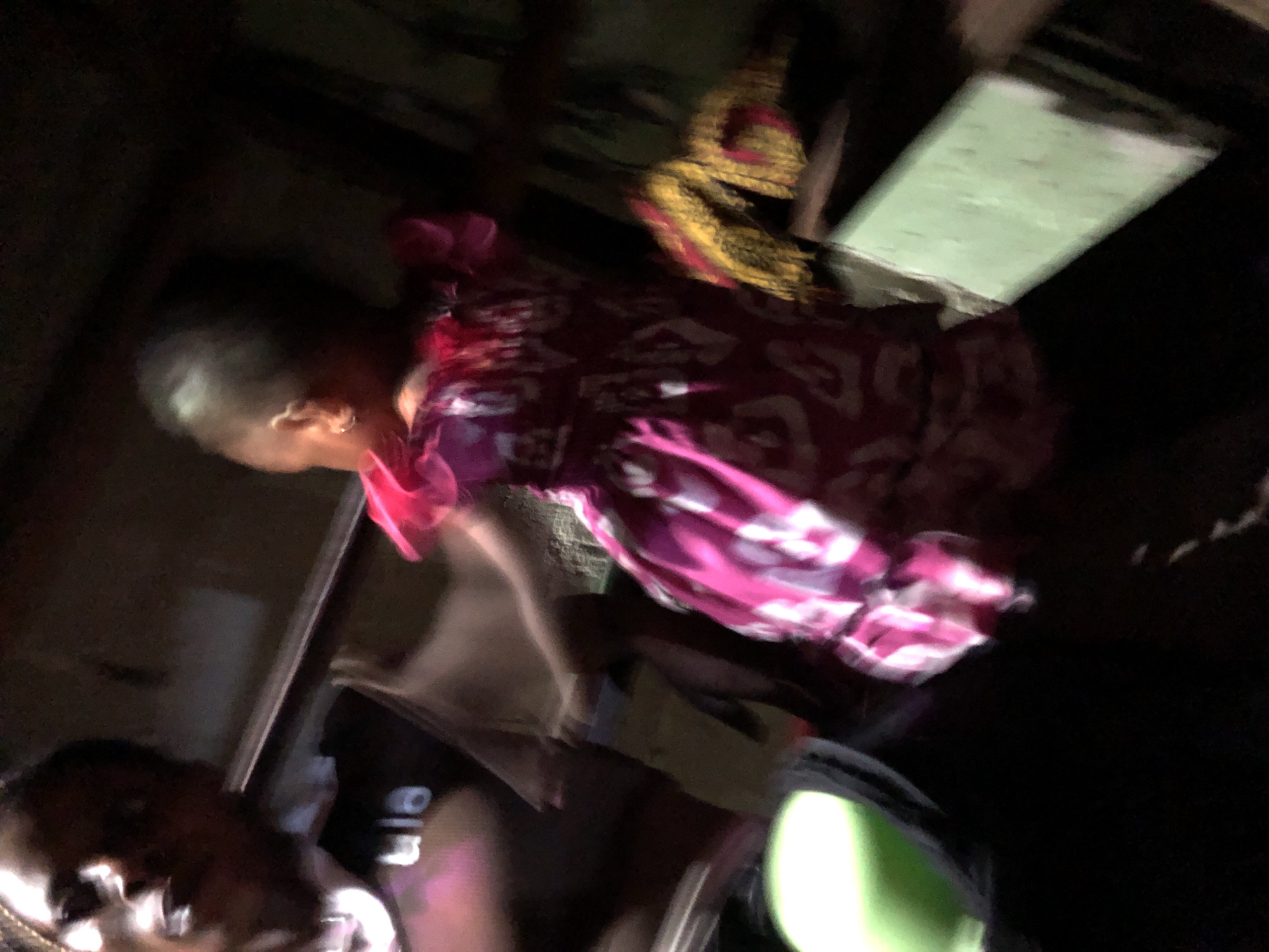 A low-angle, blurry photograph taken indoors shows a child wearing a patterned pink and white dress. The child is facing to the left of the frame, with their head near the upper left corner. The environment is dark, but a light-colored rectangular object can be seen near the top right corner. Other people and objects are visible, but not clearly discernible due to the blurriness of the image. The location is stated as Moba, Nigeria.