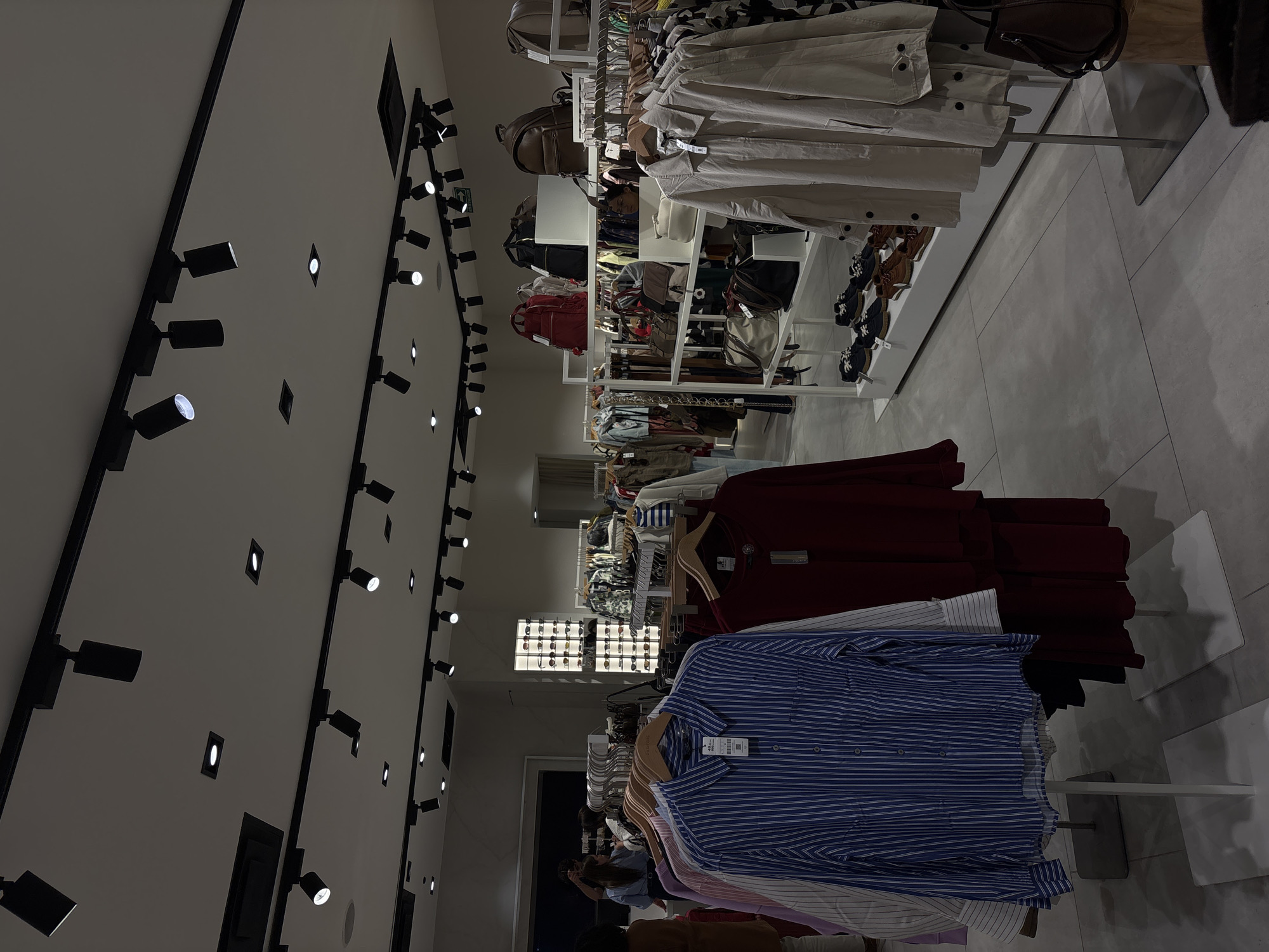 The image shows the interior of a clothing store. On the left, there is a white wall adorned with a series of black track lights. Clothing racks are filled with various garments, including shirts and jackets in different colors and styles. Some clothing items, such as shoes and bags, are displayed on shelves. In the background, there's a window display of sunglasses. People are visible in the background. The location is in the city of Puebla, Mexico.