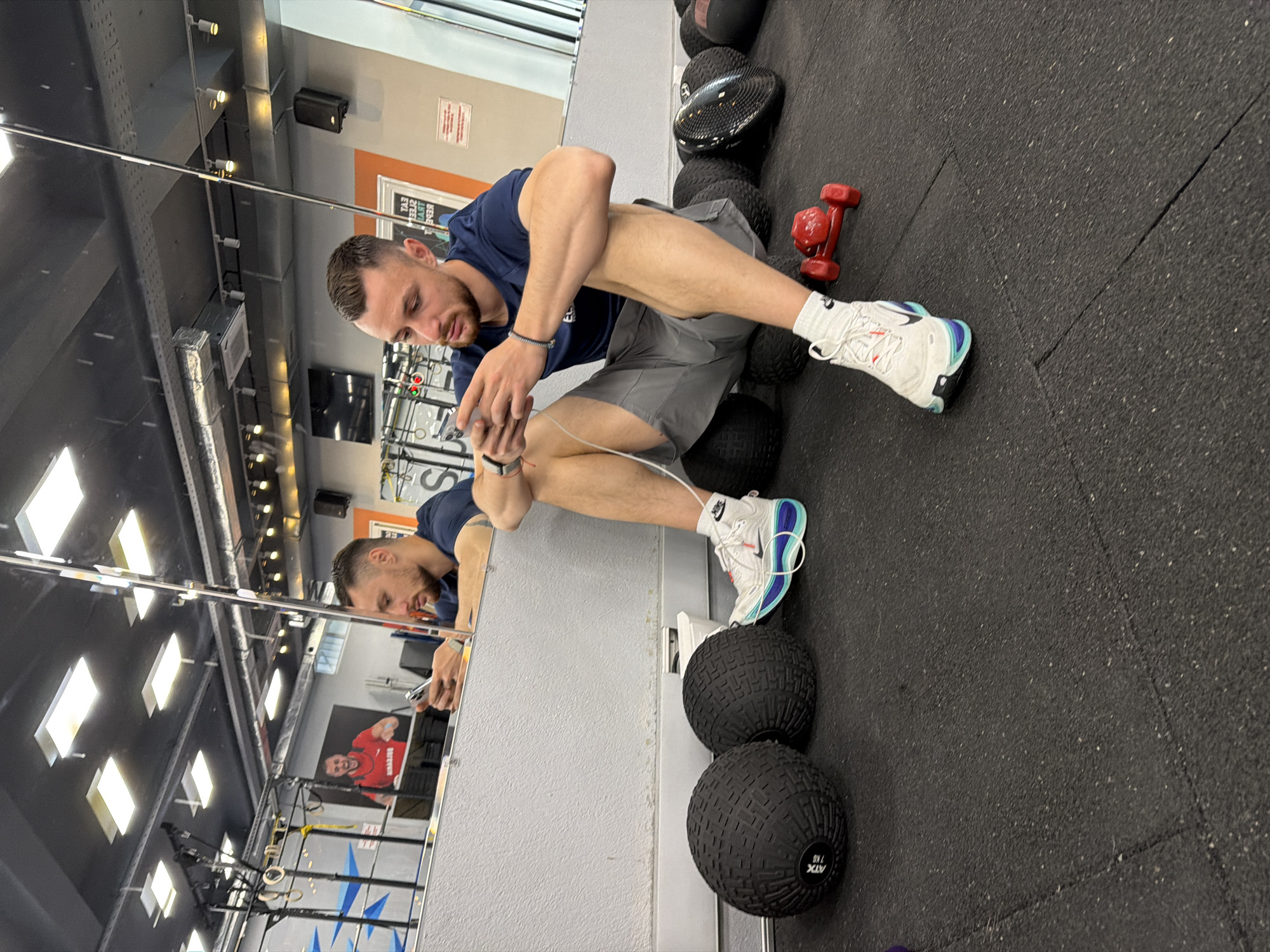 A man is sitting on a stack of medicine balls in a gym, looking at his phone. He is wearing a dark blue t-shirt, gray shorts, and white sneakers with blue and black accents. Next to the medicine balls is a red dumbbell. The background includes a mirror reflecting the gym equipment, posters, and lights. The gym appears to be located in the city of Sofia, Bulgaria.