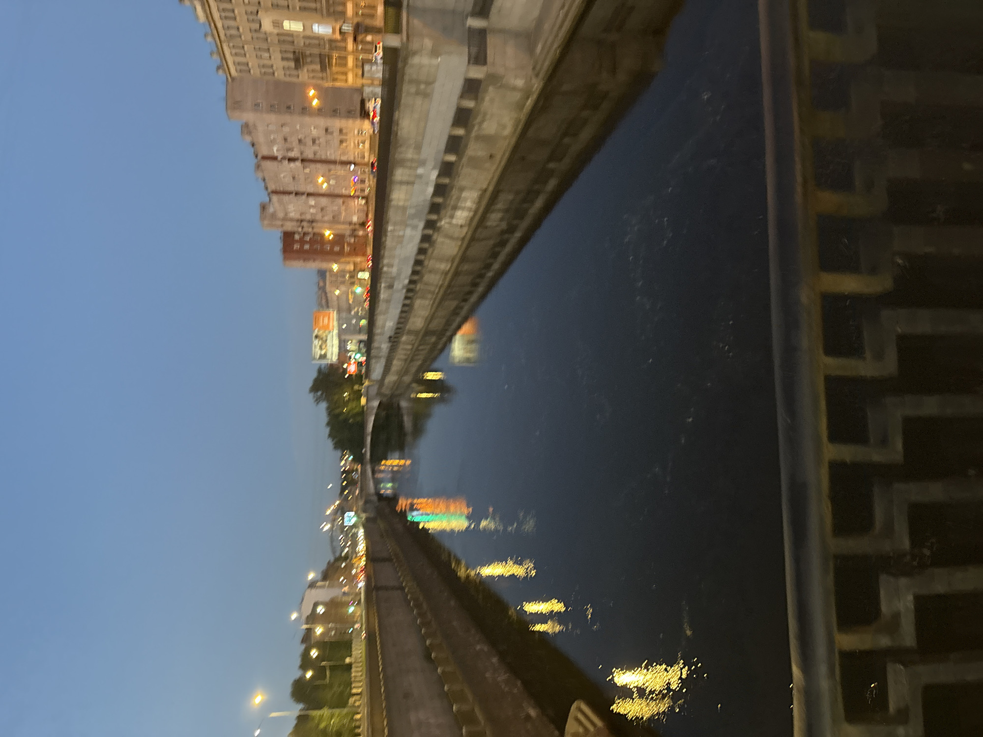 The image depicts a canal in St. Petersburg, Russia, at dusk. The canal is flanked by retaining walls, with buildings lining one side of the canal. Lights from the city are reflected in the dark water. A railing is visible in the foreground, suggesting the vantage point is from a bridge or embankment. The sky is a clear, pale blue, indicative of twilight. The buildings are of a typical European architectural style, with multiple stories and windows. The overall impression is a serene urban landscape illuminated by artificial lights.