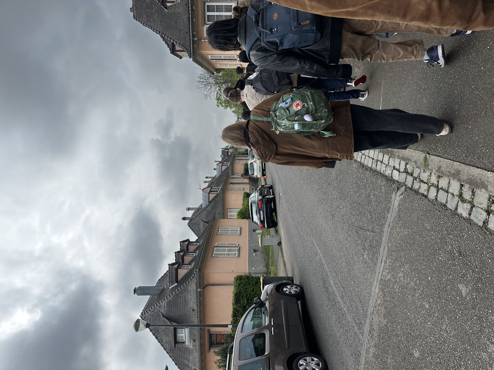 A street in Strasbourg, France, shows a group of pedestrians walking on the sidewalk. The street is lined with peach-colored houses with dark tiled roofs. Several cars are parked along the street. The sky is overcast. The pedestrians are wearing casual clothing, and some have backpacks. The sidewalk is paved with grey stone.