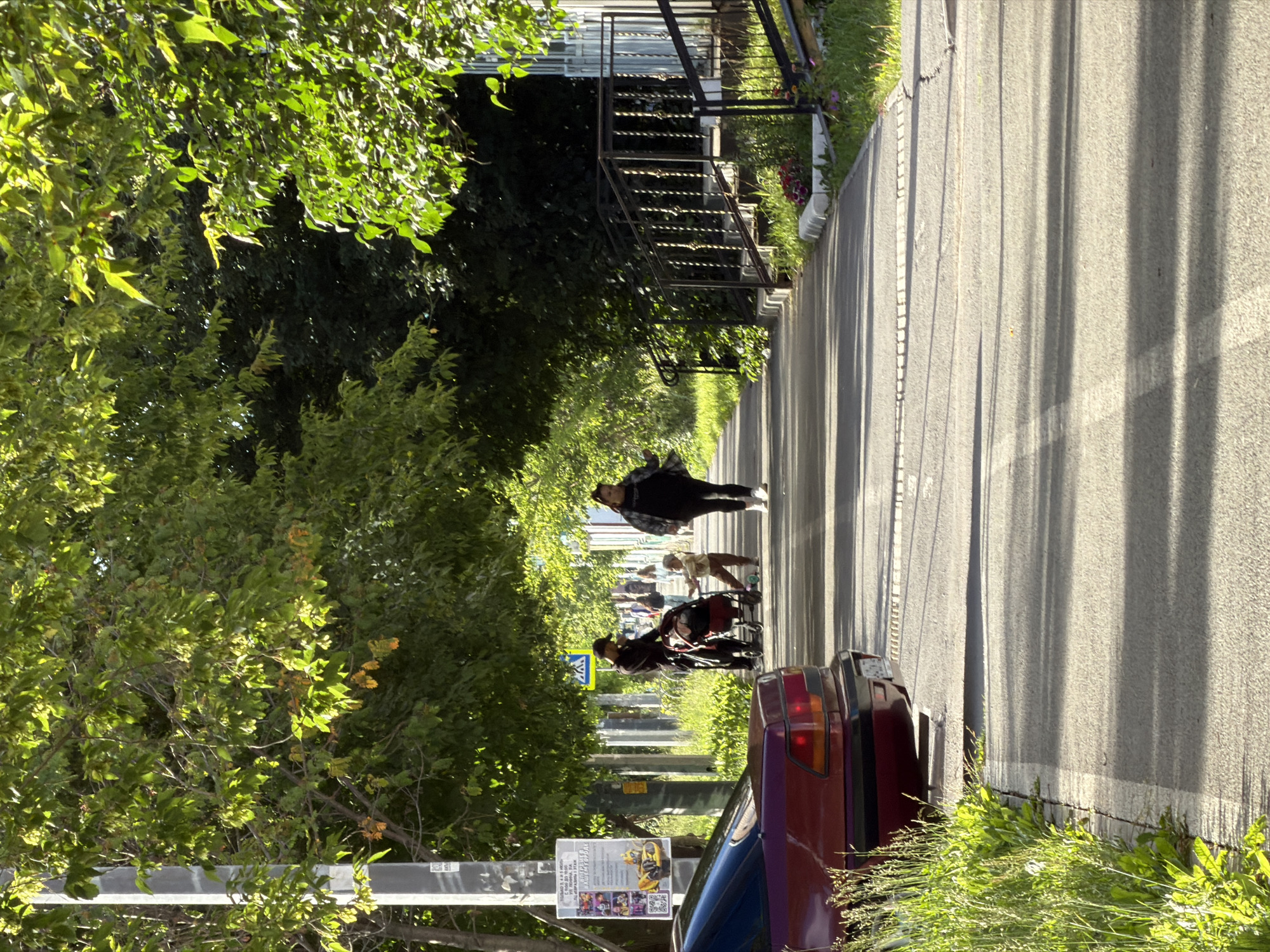 This image depicts a street scene with several people and objects. In the foreground, a red car is parked on the right side of the frame, with its taillights visible. To the left of the car, a street sign is mounted on a pole. Beyond the car, a paved walkway or road stretches into the distance, lined with trees and vegetation.
Several individuals are visible walking or standing along the walkway. One person, wearing a dark jacket and pants, is walking away from the camera. Further down the walkway, another person is seen pushing a stroller or cart. Two more figures are further in the background, one of whom appears to be on a skateboard.
The scene is bathed in sunlight, casting long shadows across the walkway, suggesting it is either morning or late afternoon. The trees on the left are lush and green, indicating a temperate season. The overall atmosphere appears to be casual and everyday.
A poster is affixed to the street sign pole, featuring a colorful image of what appears to be a racing vehicle and several small figures. Some text is visible on the poster, but it is too small to be deciphered. A pedestrian crossing sign is also visible on another pole. The setting seems to be a residential or urban area with sidewalks and greenery.