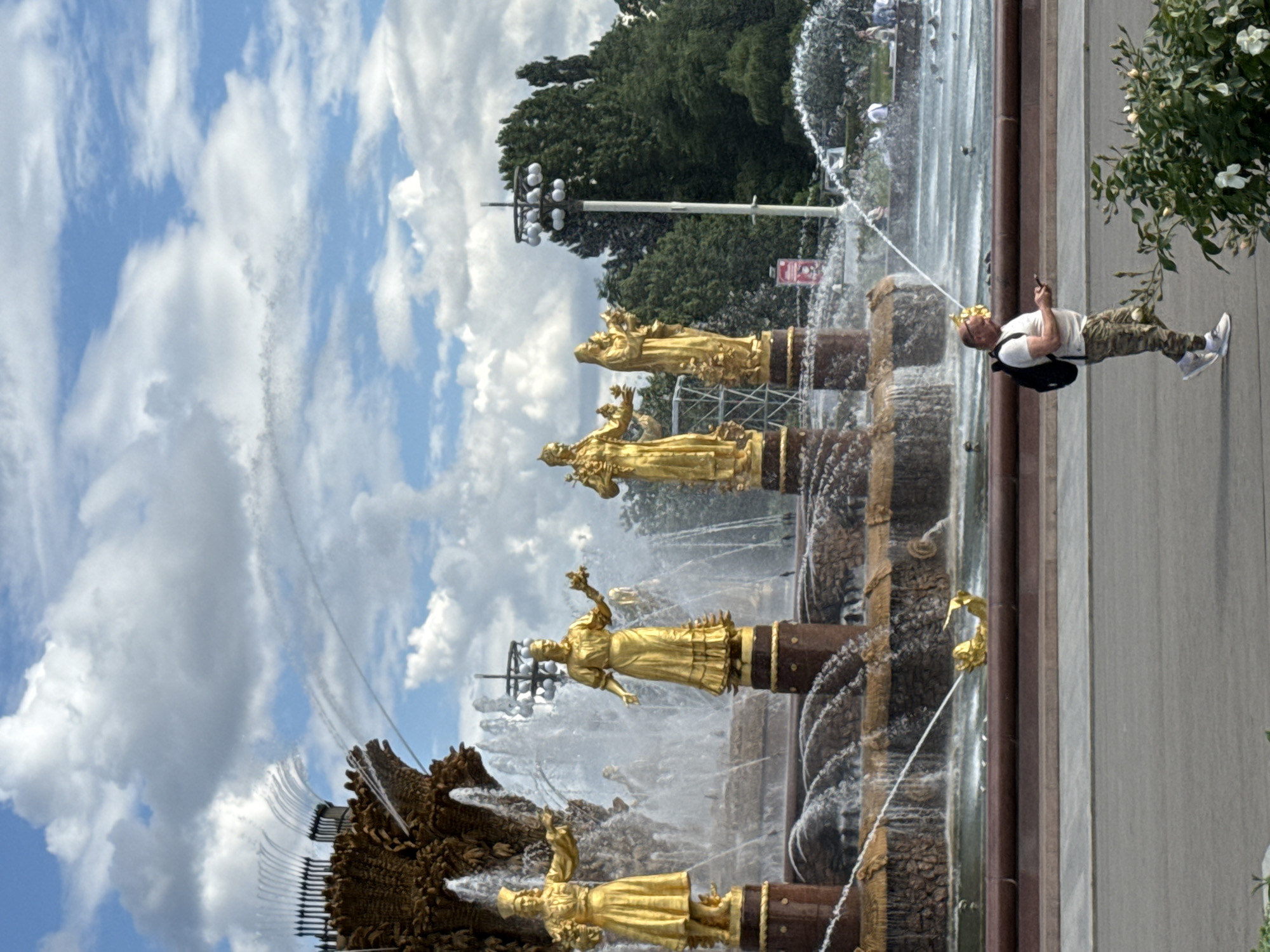 This media file depicts a man standing near a large, ornate fountain in Moscow, Russia. The man is wearing a white t-shirt, camouflage pants, a backpack, and a golden crown. He is holding a mobile phone and appears to be taking a picture or video of the fountain. The fountain itself is a central feature, with multiple tiers adorned with golden statues of figures. Water is actively spraying from various points of the fountain, creating a dynamic and refreshing atmosphere. The sky above is blue with scattered white clouds, suggesting it is a pleasant day, likely daytime. Lush green trees are visible in the background, indicating a park or public garden setting. A lamppost with multiple lights stands near the fountain, and there is a small red sign on a pole in the background, though the text on it is not legible. The overall scene suggests a tourist or local enjoying a summer day at a significant landmark. The man's attire and the crown he's wearing might indicate a playful or festive mood, possibly inspired by the golden statues or the grandeur of the fountain.