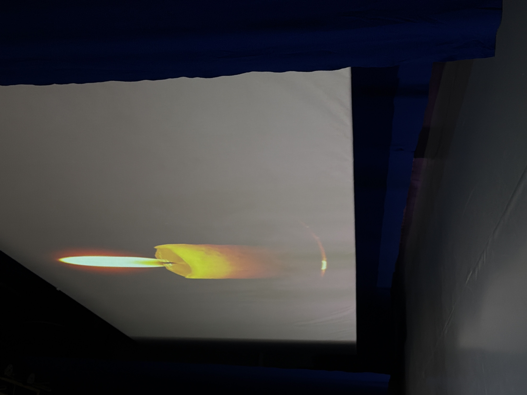 The image shows a projection of a candle flame on a screen, likely inside a building. The screen is framed by dark blue fabric or material along the top and sides. To the right of the screen is a grey wall. The candle image appears to be positioned in the lower center of the screen. The location, Perm, Russia, is not evident in the image.