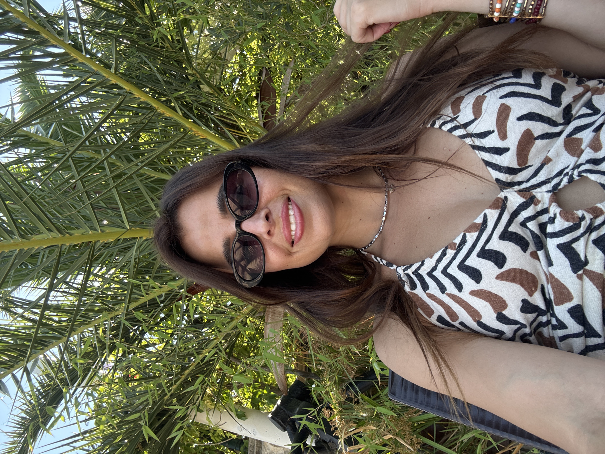 A woman is lying down outdoors, possibly in a park or garden, surrounded by lush green foliage and palm leaves. She is captured in a close-up shot, with her head tilted back and her hair fanned out. She is wearing sunglasses, a patterned top with a zebra-like print, and a silver necklace. On her wrist, she has several colorful bracelets. The woman is smiling, revealing her teeth, and her eyes are visible behind her sunglasses. The sunlight casts a warm glow on her face, suggesting it is daytime. The greenery surrounding her indicates a natural setting, possibly a sunny day. The composition of the image and the relaxed pose suggest a candid moment, perhaps a selfie taken while enjoying the outdoors. There is no visible text in the image.