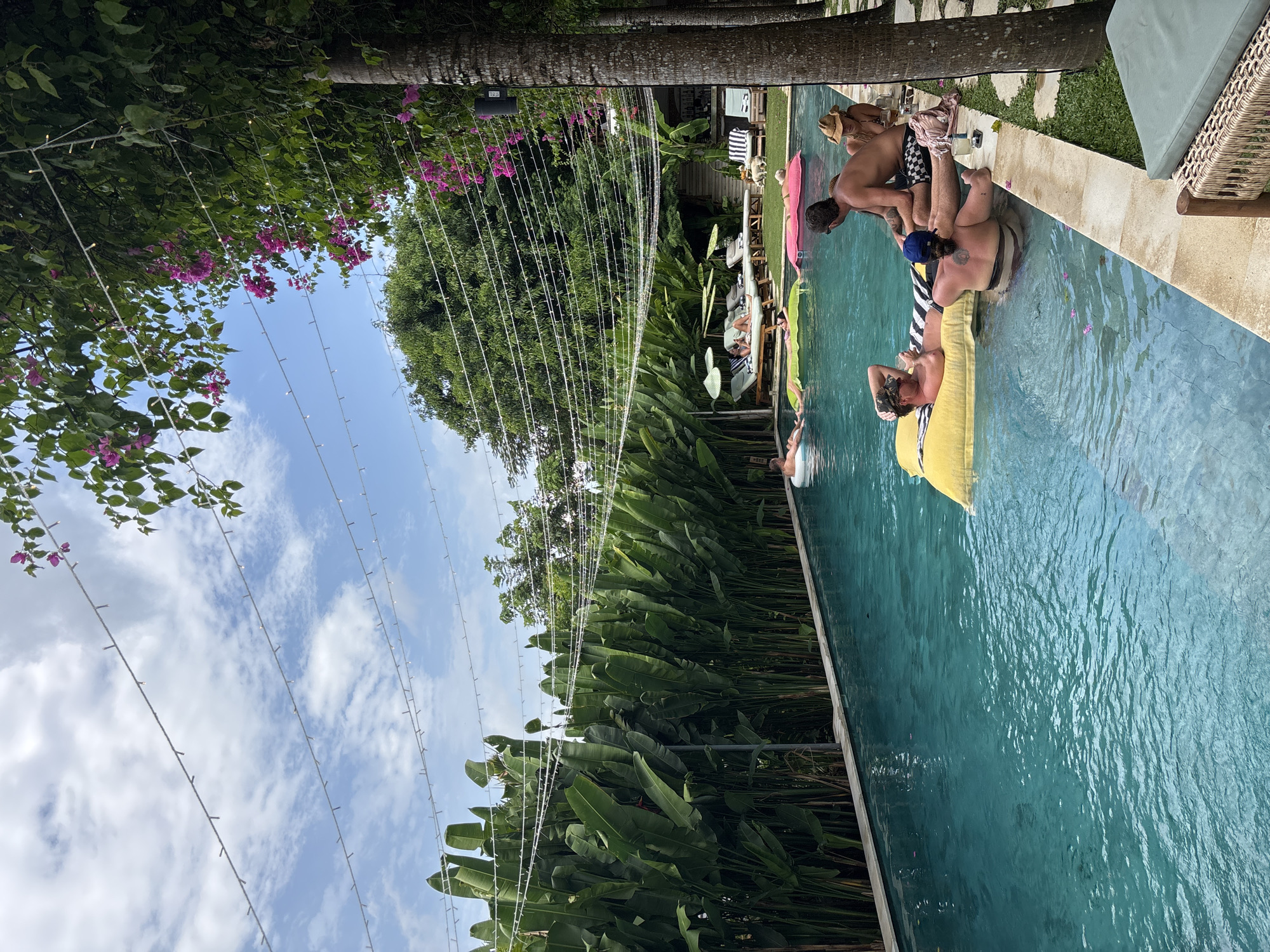 The scene shows a group of people enjoying a sunny day at a swimming pool, potentially in Kuta Selatan, Indonesia.
**Setting:** A clear, blue swimming pool is the primary element. Tropical vegetation surrounds the pool, including lush green plants and trees. String lights are suspended overhead, creating a festive atmosphere. The sky is partly cloudy.
**People:** Several individuals are present.
* A person is floating on a yellow inflatable raft in the pool.
* Two individuals appear to be interacting, with one possibly assisting the other into or out of the pool. One of the men wears checkerboard shorts.
* Other people can be seen lounging near the pool.
**Objects:**
* Various lounge chairs and sunbathing mats are placed around the pool.
* A striped towel is visible on one of the chairs.
* Beverages are placed near the edge of the pool.
**Actions and Interactions:**
* People are relaxing and socializing around the pool.
* Some are actively engaging in swimming or assisting others.
**Overall Impression:** The image depicts a casual, leisure scene with people enjoying a pool setting in a tropical location.