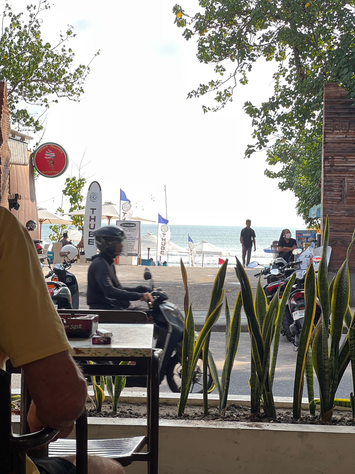 In Kuta, Indonesia, a view from a cafe's outdoor seating area shows a street scene with the ocean in the background. In the foreground, a person in a yellow shirt is partially visible, seated, with their arm resting on a dark metal chair. On a table in front of them is an ashtray with cigarettes and a lighter, as well as a pack of Camel cigarettes. To the right, tall, leafy snake plants are growing in a planter. Beyond the planter, a man wearing a black shirt and pants walks on a beach. To the left of the man, two people are seated in chairs near an umbrella. Further left, several motorcycles are parked. A person on a black motorcycle wearing a black helmet and jacket rides past. A sign for "Gelato Massimo" is visible on a brick wall, with an image of an ice cream cone. Surfboards are propped up vertically with "THUBE" written on them. In the distance, the ocean stretches out under a bright sky.