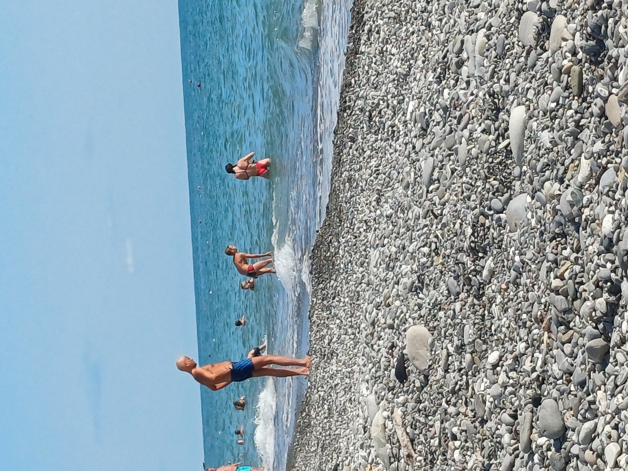 This is a vertical, eye-level, wide shot of a pebbly beach in Sochi, Russia, on a bright, sunny day. The water is a clear, vibrant blue, with small waves lapping at the shore. The beach is covered in smooth, grey and white pebbles of various sizes.
Several people are enjoying the beach and the sea. In the foreground, a man with a shaved head and blue swim trunks is standing in the shallow water, looking out towards the sea. Further back, a man in red swim trunks is standing in the water, with another person behind him. There are other figures scattered in the water and near the shore, though they are less distinct. The overall mood appears relaxed and summery, with people engaging in typical beach activities like swimming and wading.
The sun is high in the sky, casting bright light and creating reflections on the water, suggesting it is midday or early afternoon. The clear blue sky indicates fair weather. The pebbly shore and the color of the sea are characteristic of the Black Sea coast in this region. There is no visible text in the image.