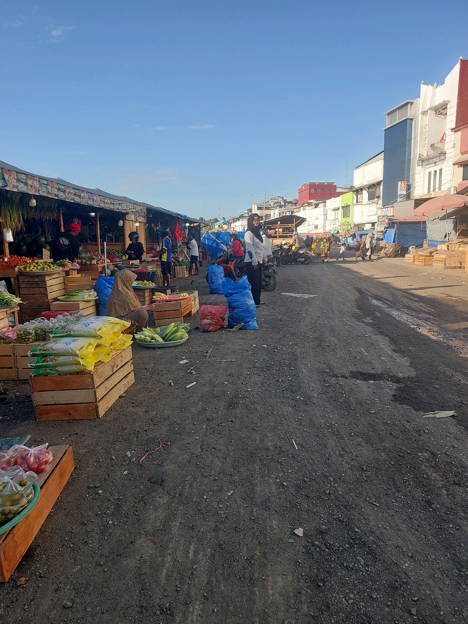 The image depicts a marketplace scene in Ambon, Indonesia, likely in a commercial or semi-commercial district given the presence of multi-story buildings in the background. The foreground shows a rough, gravelly surface, indicative of an outdoor market or a less-developed street. Several market stalls are visible, featuring a variety of produce displayed in wooden crates and bags.
In the left and center of the image, market vendors and customers are engaged in transactions or preparing goods. One woman, wearing a headscarf and patterned clothing, is seated near a display of corn and other vegetables. Several sacks, some blue and some red, are scattered around the area. Bright yellow bags, possibly containing food items or supplies, are stacked in wooden crates on the left. Further back, more market stalls are lined up, with awnings providing shade.
In the background, a street stretches into the distance, with various buildings lining its sides. Some of these buildings appear to be shops or commercial establishments, with one prominent building featuring a modern glass facade and signage. Vehicles, including a yellow three-wheeled motor vehicle (likely an auto-rickshaw or "becak motor") and motorcycles, are present on the street, suggesting ongoing activity and transportation. The sky is clear and blue, indicating daylight. The overall atmosphere is that of a busy, functional marketplace in an urban setting.