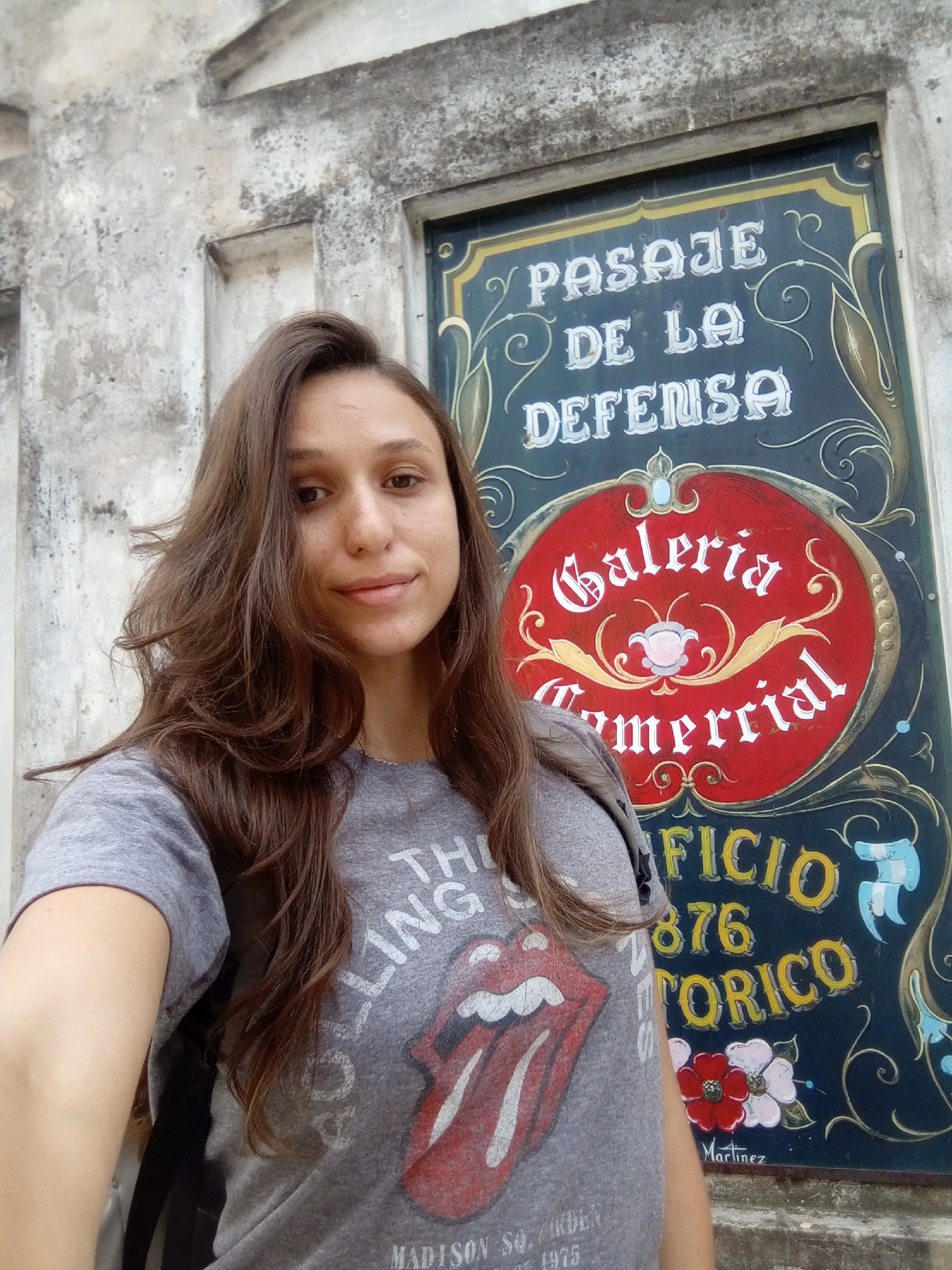 A woman stands in front of a sign in Buenos Aires, Argentina. The sign reads "Pasaje de la Defensa, Galeria Comercial, Edificio Historico 876" and has floral designs. The woman wears a grey t-shirt with a Rolling Stones tongue logo. The building behind the sign appears old and weathered. "Martinez" is written in the bottom right corner of the sign. The t-shirt has "MADISON SO. GARDEN" and "1975" written at the bottom.